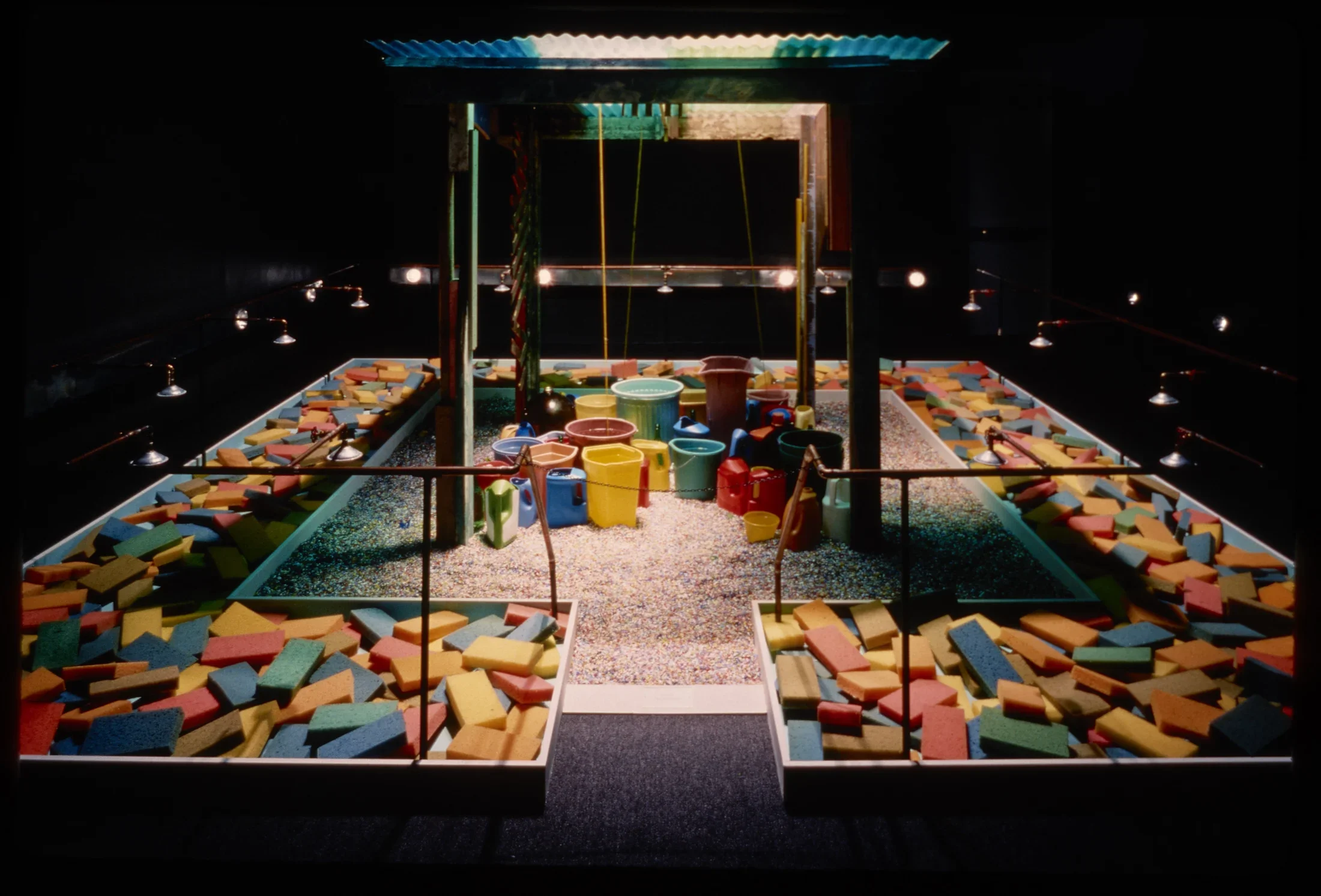 Wooden playset with spotlight on plastic sand castle buckets in sand, surrounded by troughs filled with colorful wooden bricks. 