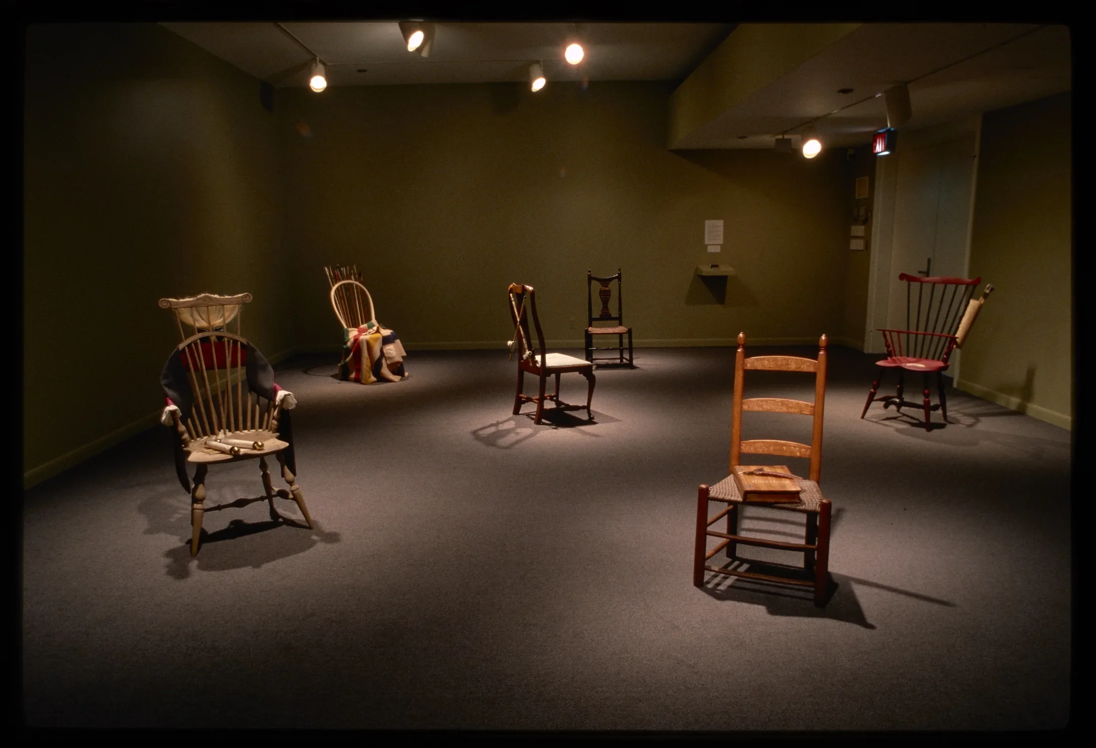 Dim room with spotlights on six spaced out colorful 17th and 18th century chairs with household items sitting on them. 