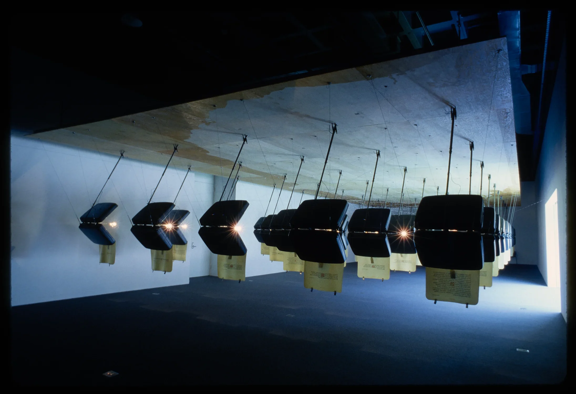 A rear view of identical hanging suitcases. Lights shine through the openings while natural light streams into the gallery. 