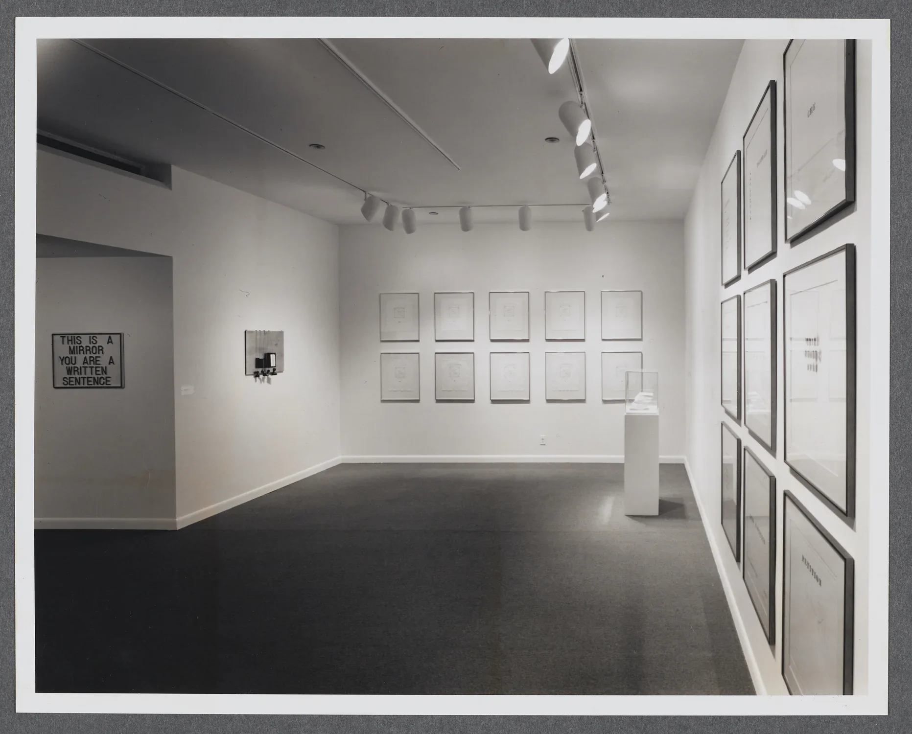 Framed works line the gallery wall in different configurations. A pedestal holding a small sculpture sits in the corner. 
