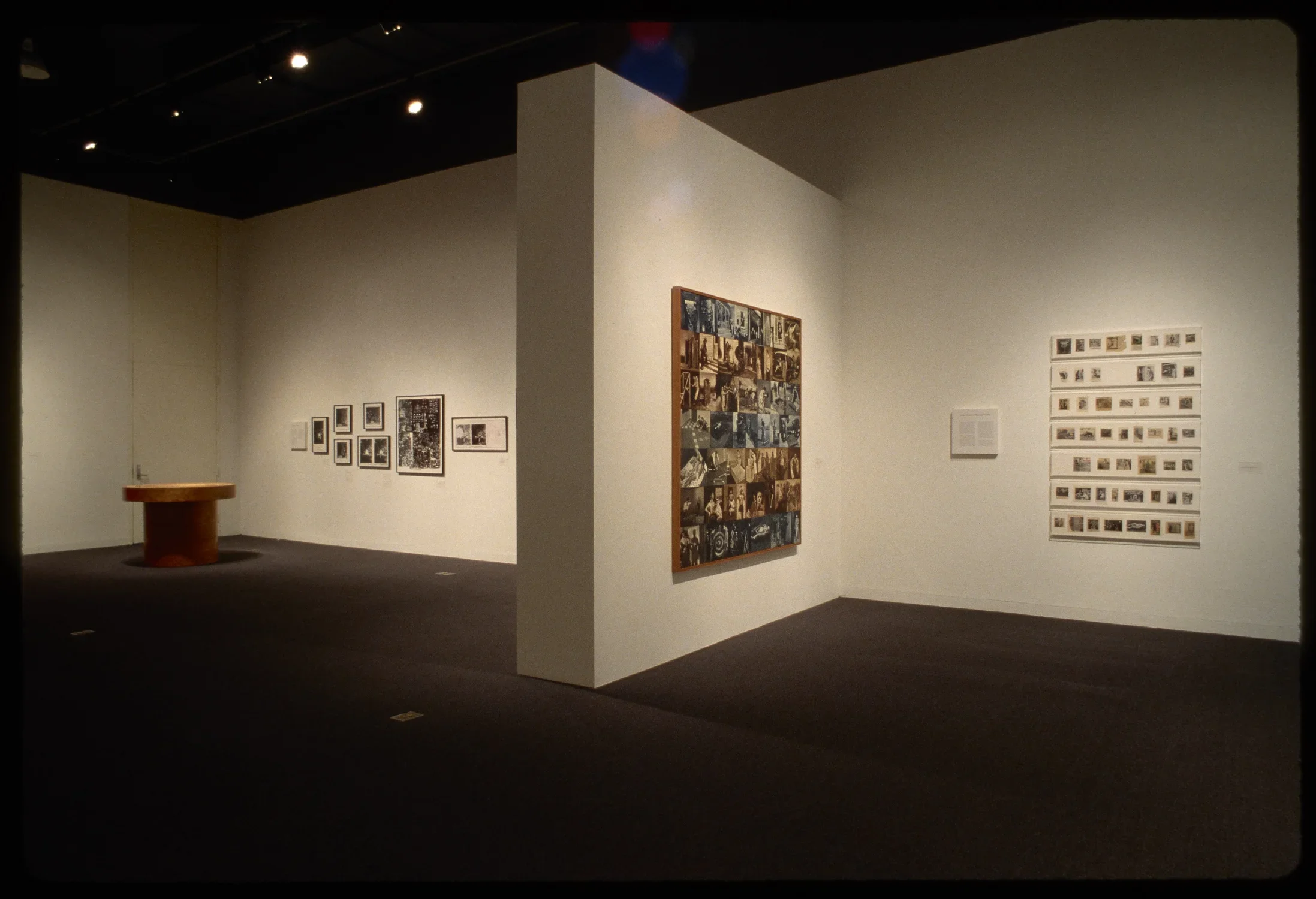 Wall separates art spaces: one with patchwork sepia photos and fifty photos, other with eight framed photos and wooden table. 