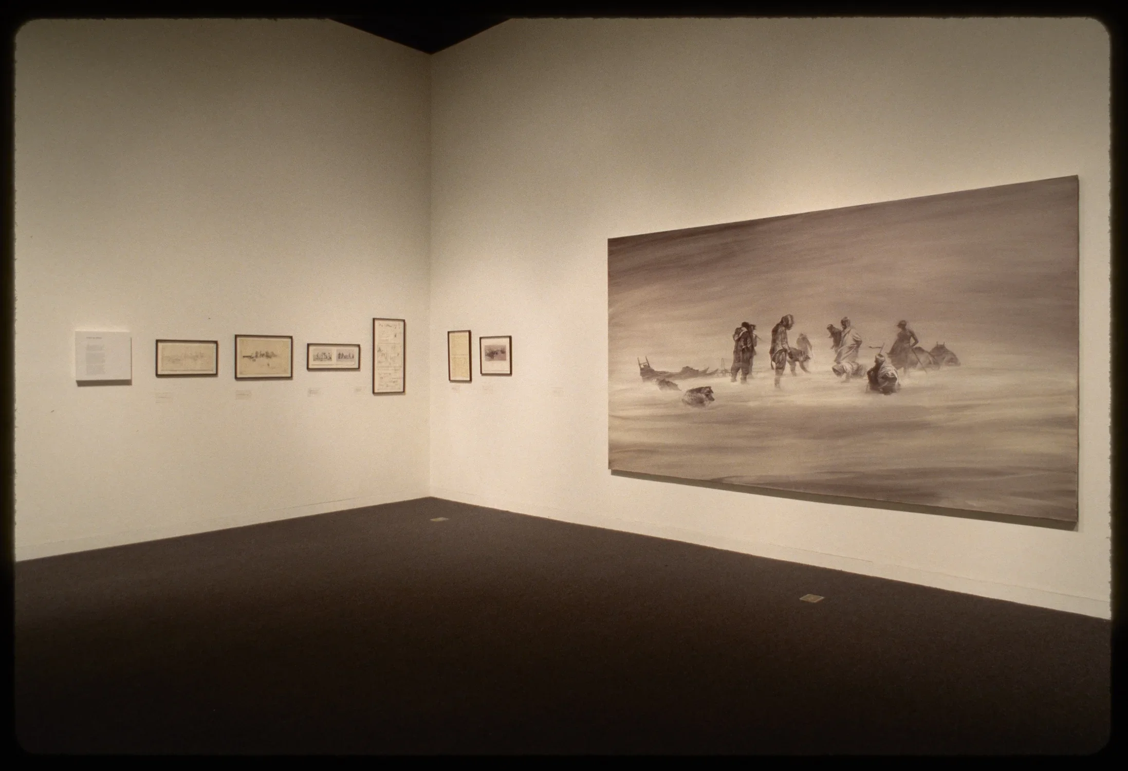 Wall text and six framed news clippings hang adjacent to large monochrome oil painting of nomads in windy desert. 