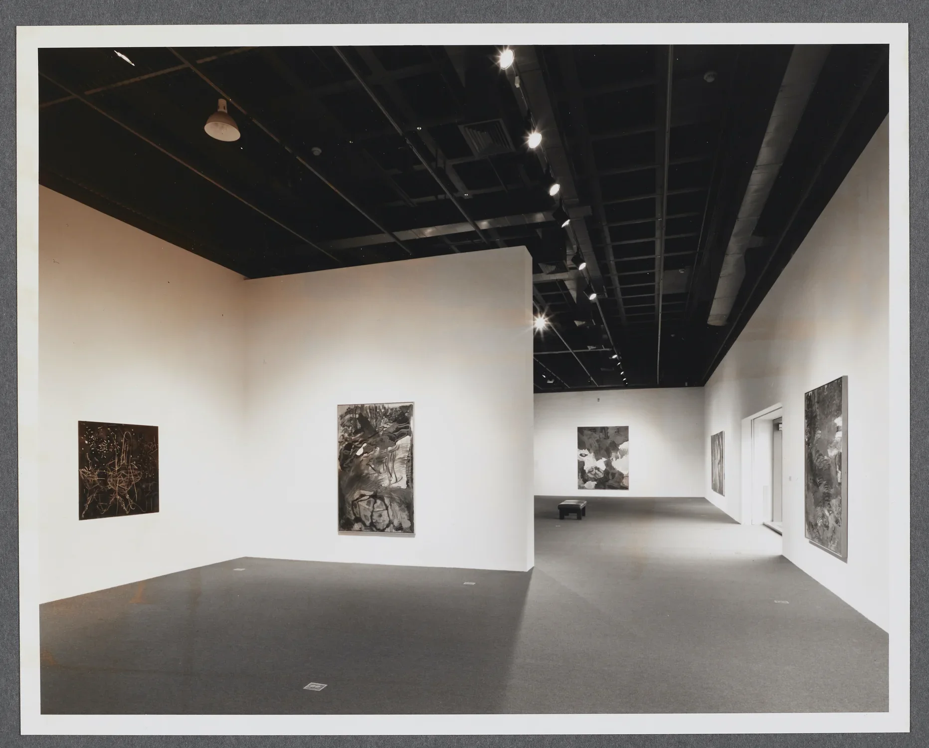 Large abstract paintings line the gallery walls as a bench sits in the center of the gallery. 