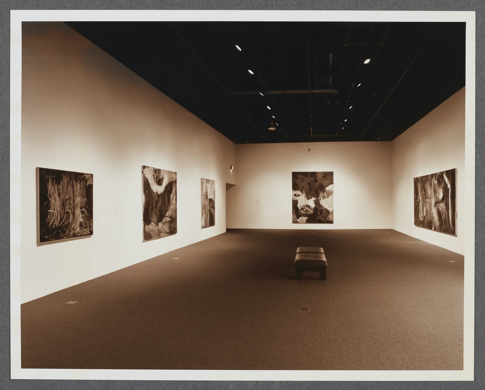 Large abstract paintings line the gallery walls as a bench sits in the center of the gallery. 