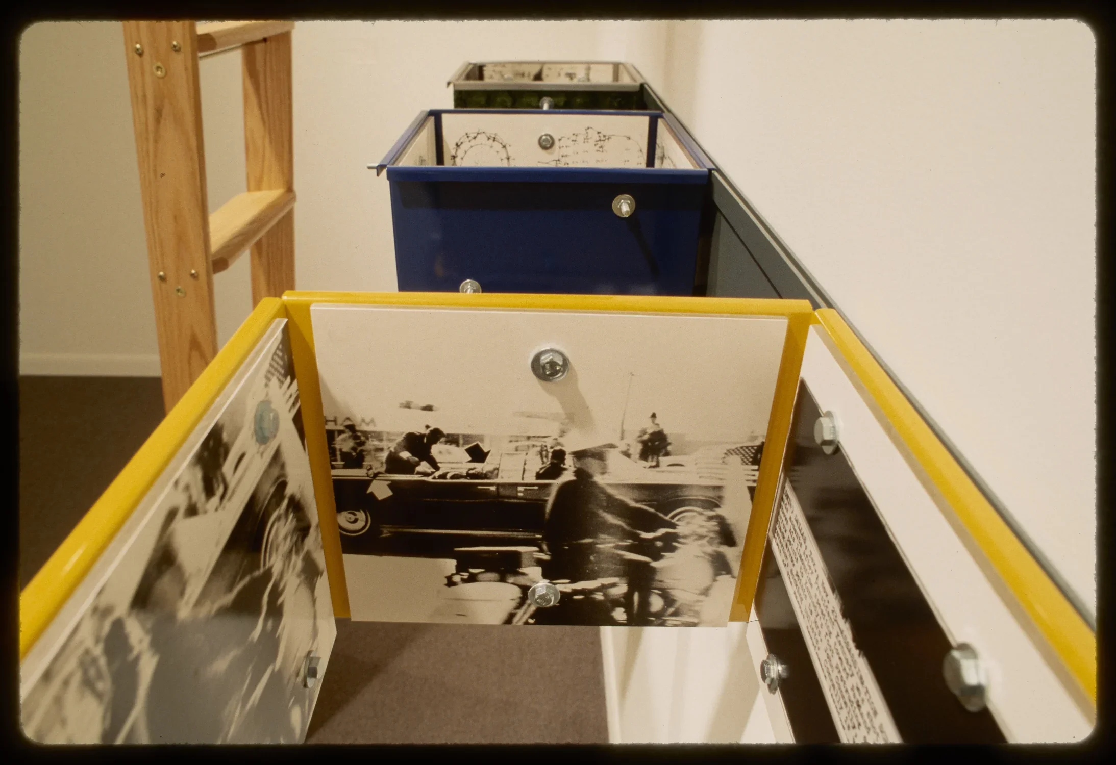 Yellow, blue, and green square boxes with no top or bottom hang on gallery wall with black and white photos lining the inside. 