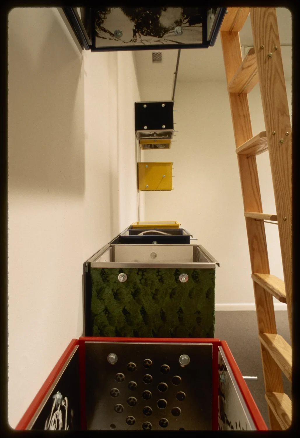 Eight colorful boxes without tops or bottoms are lined on the inside with metal sheets with holes and white and black photos. 