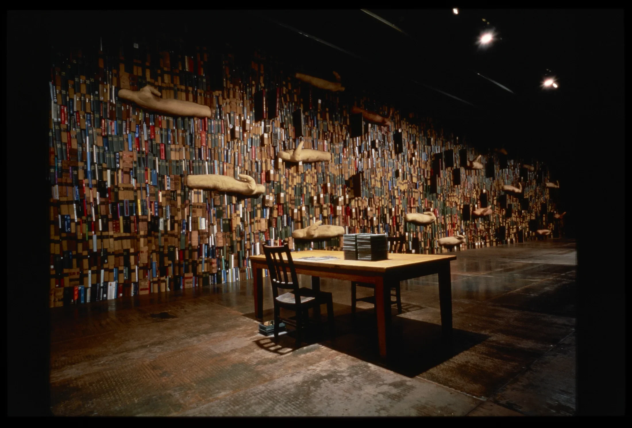 Wall is lined with 20,000 colorful books with thirteen androgynous bulbous “bodies,” table and chair sits in middle of room. 