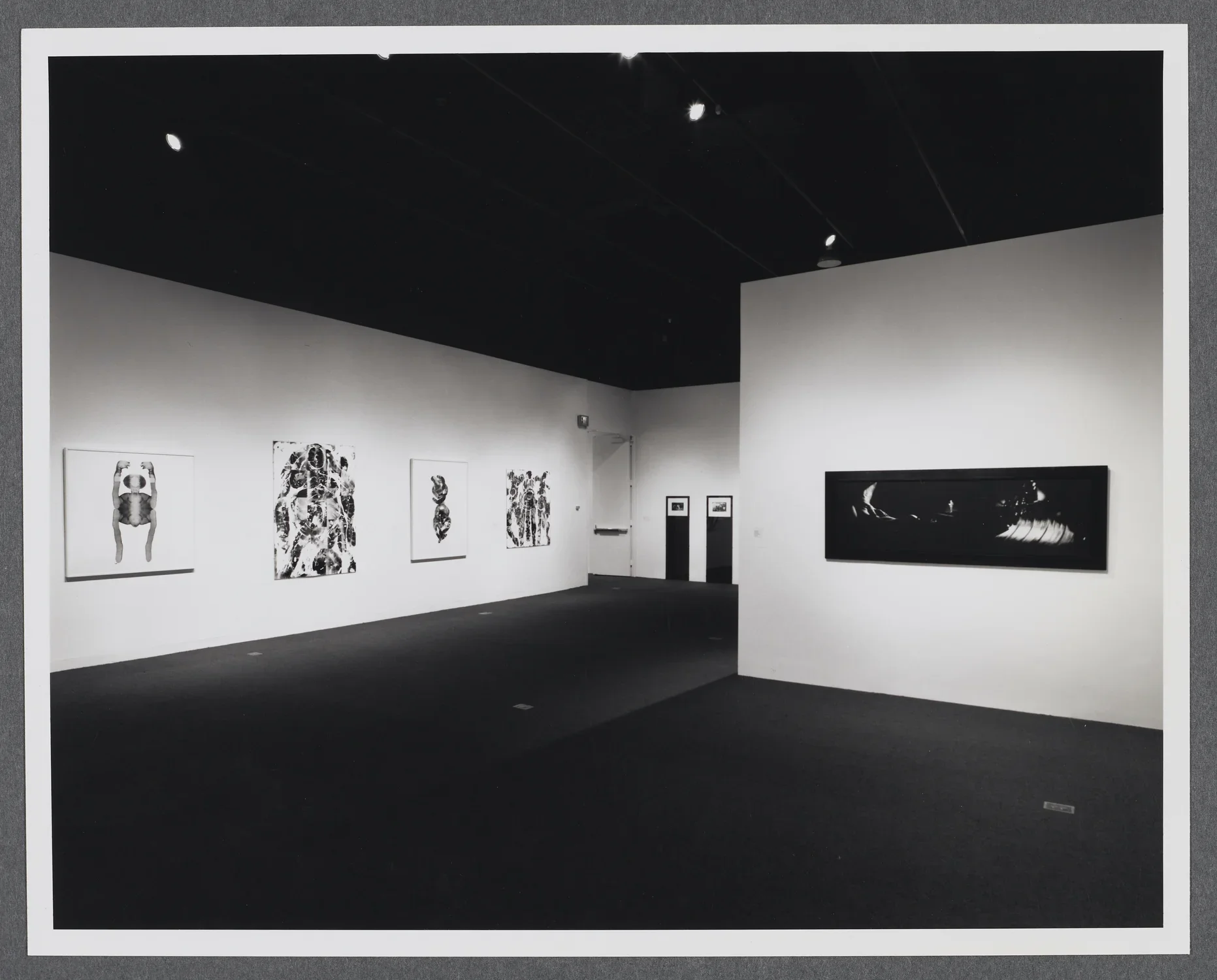 Large abstract artworks hang around the gallery space. A gallery wall stands in the center of the room.  