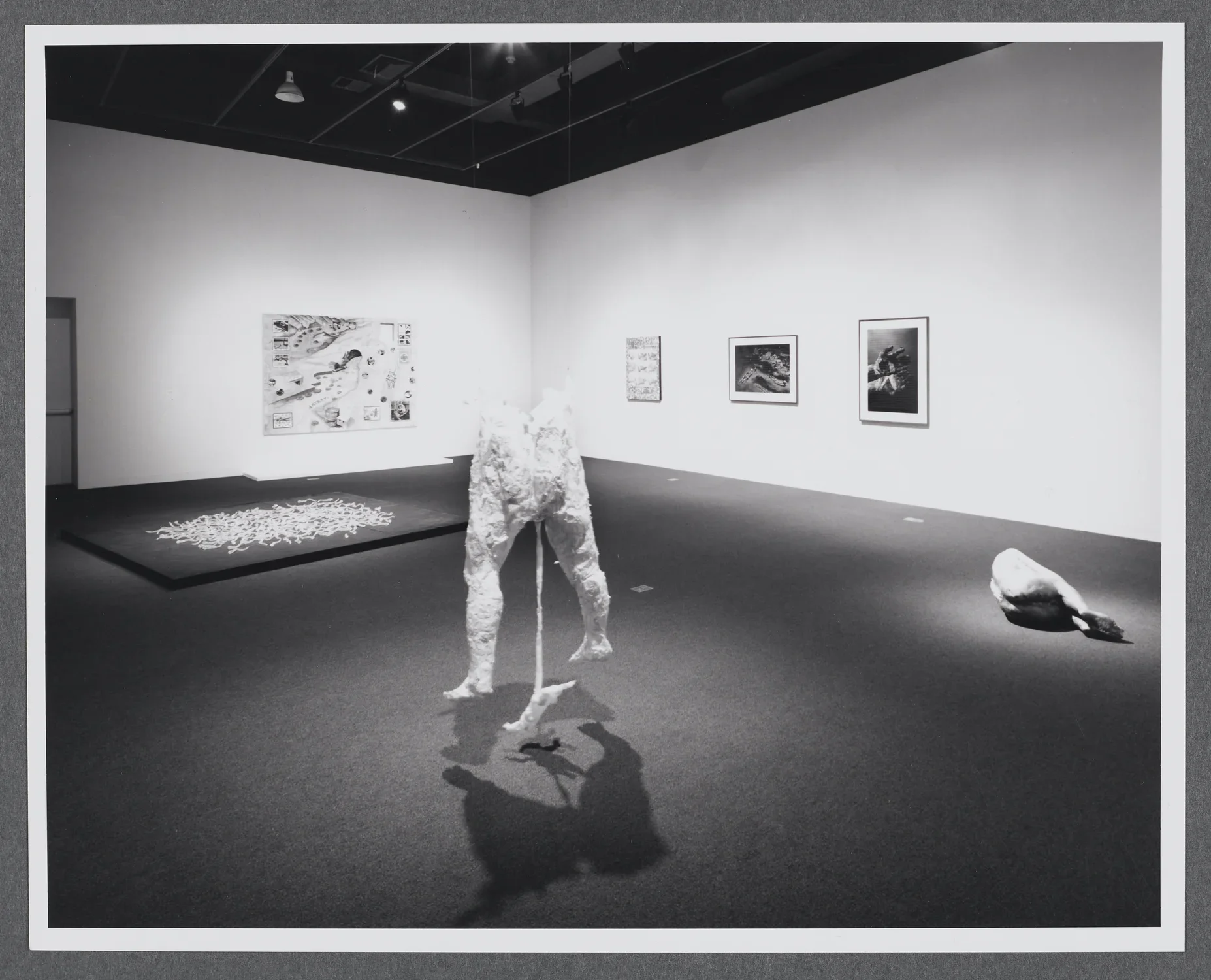 A sculpture of two standing legs is placed in the center of the room. Spotlight from above, the sculpture casts two shadows. 