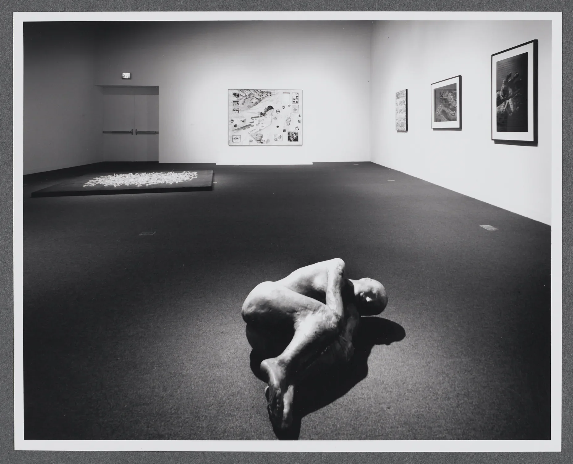 A sculpture of a human figure is curled up on its side. It sits completely alone compared to the other works in the room. 