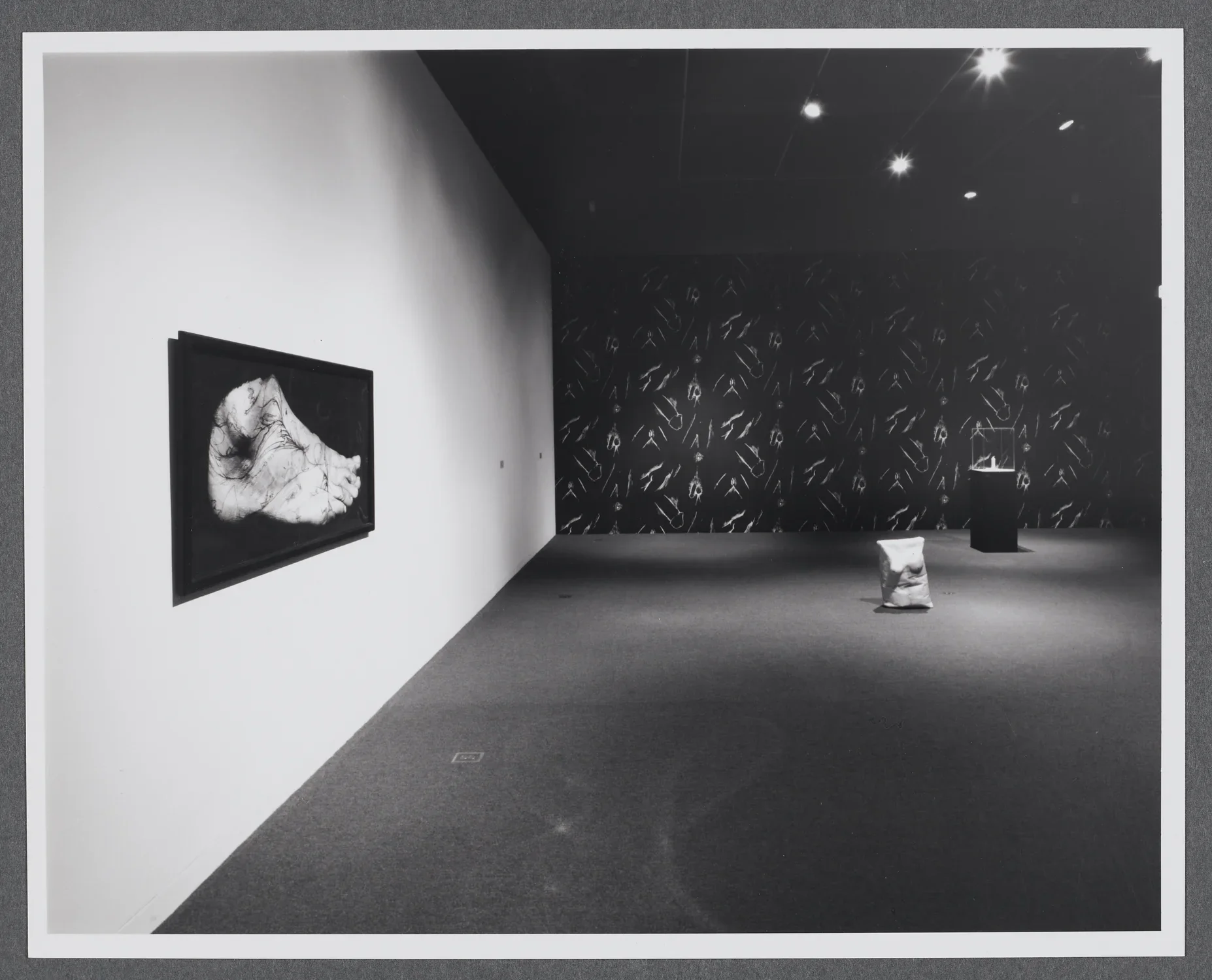 An image of a human foot is hung on the wall. In the distance is a sculpture of a sack made to look like a female torso.  