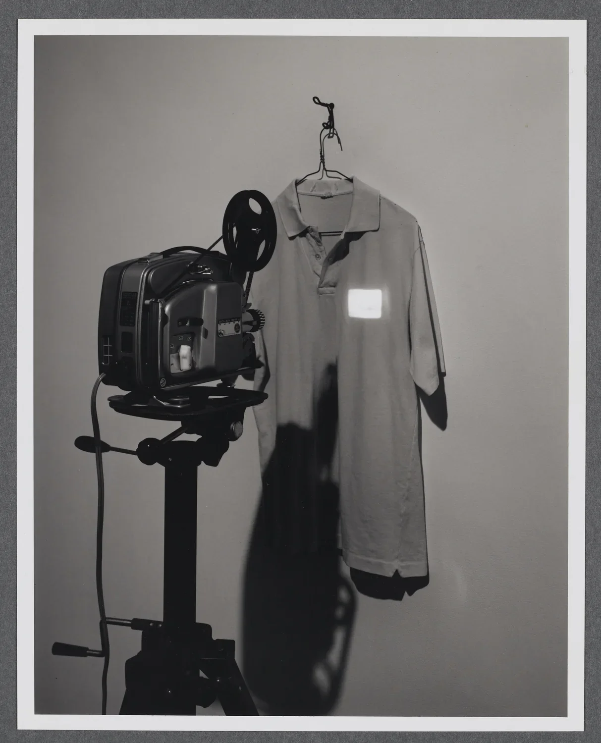 A collared shirt hangs on the gallery wall. A projector projects a tiny image onto the upper right hand side of the shirt. 