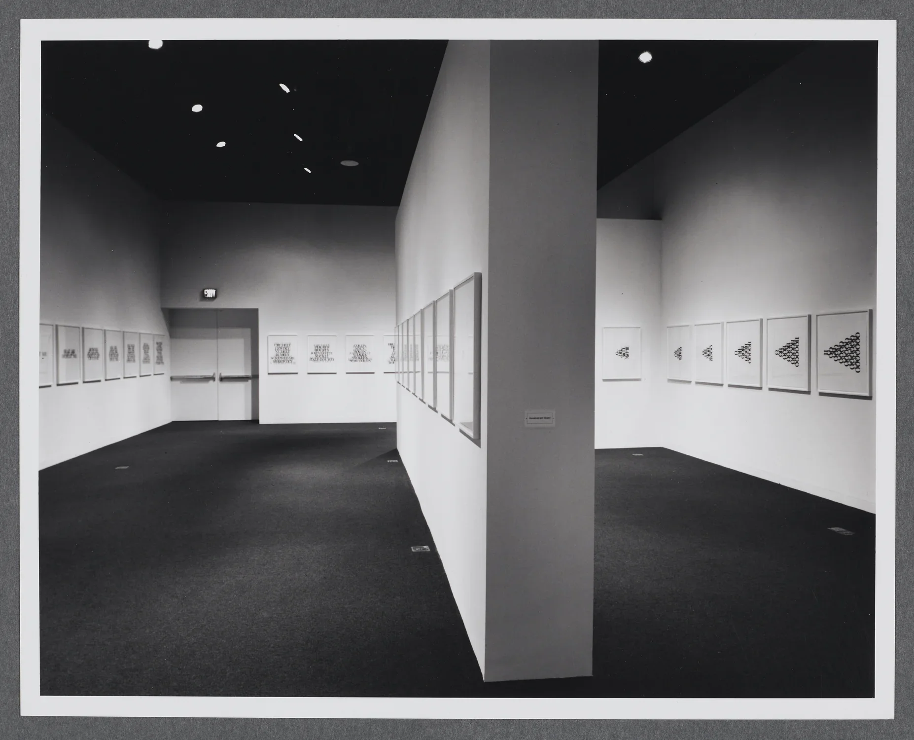 A gallery wall splits the room in half. Works of the same size are displayed in a row going across all of the gallery walls.