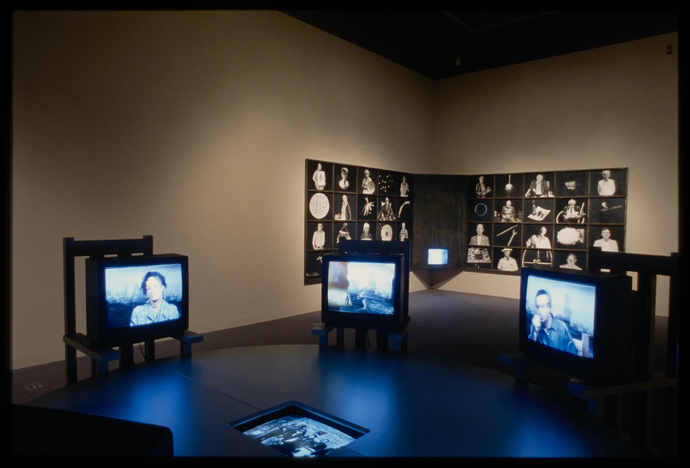 Three tvs with videos of people sit in ring in front of black and white photos of people and items covering the wall corner.