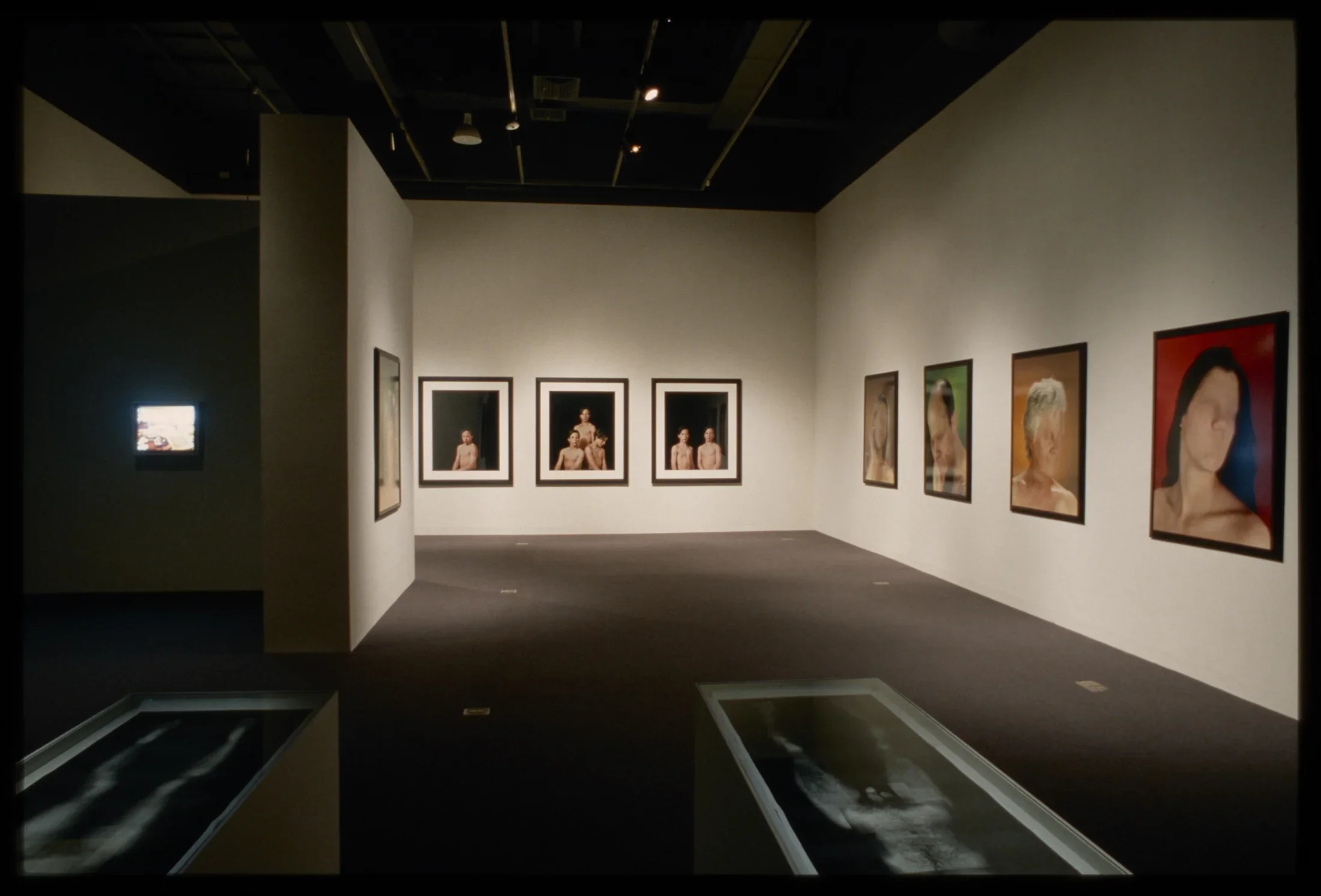 Black and white nude images sit in front of one lit screen and eight headshots of people without eyes or mouths hung on walls.