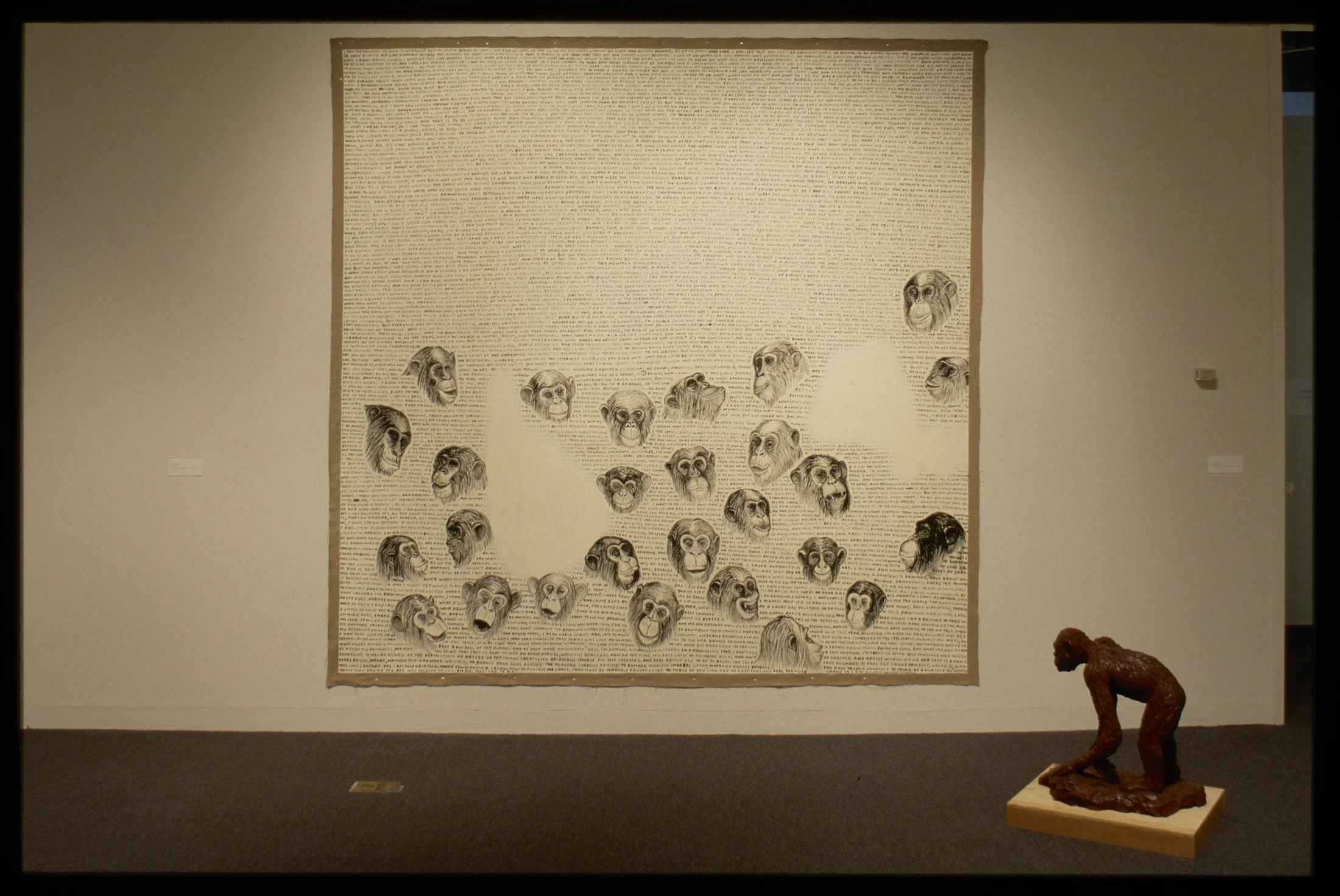 Brown clay ape sculpture stands in front of large wall mural with inked apes heads with various facial expressions and text.