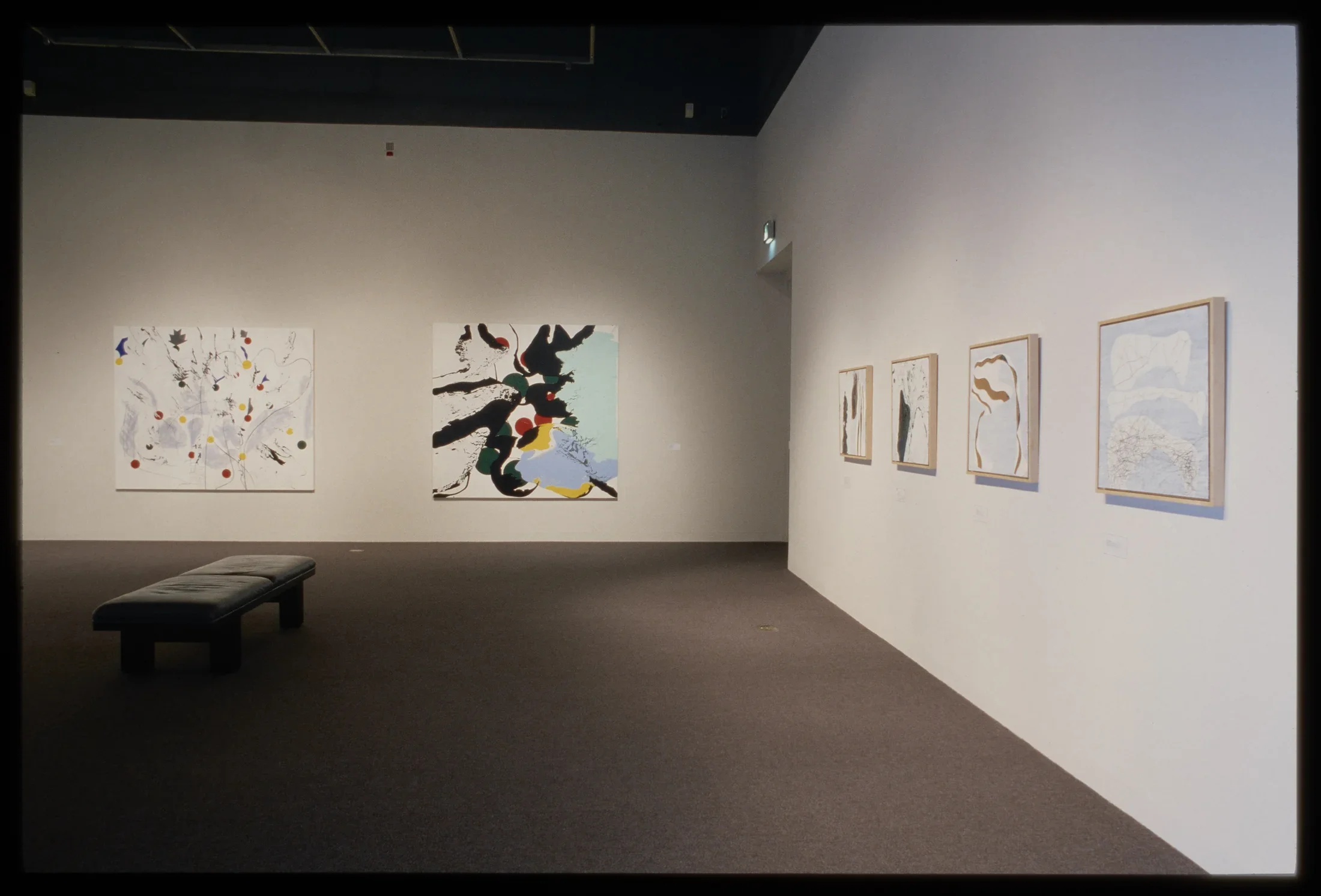 Two large and four small abstract paintings with white background and subtle colorful splotches and dots hang on adjacent walls.