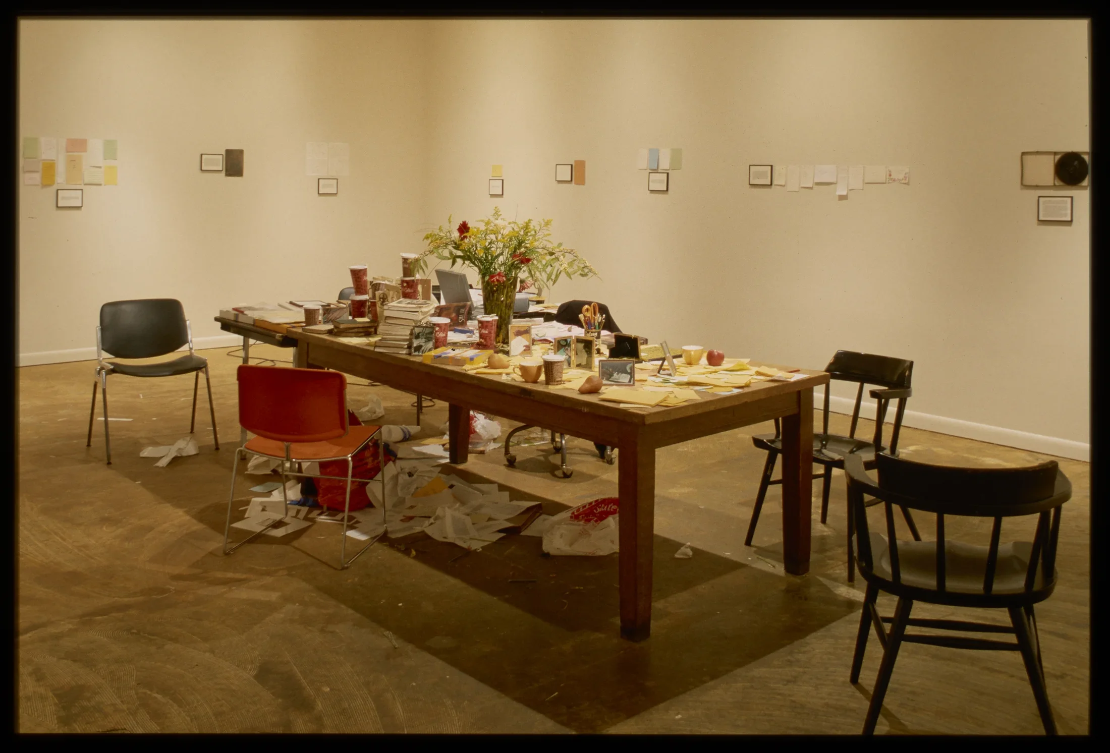 Seen at an angle, a table littered on top and below with cups and papers, surrounded by several randomly placed chairs.