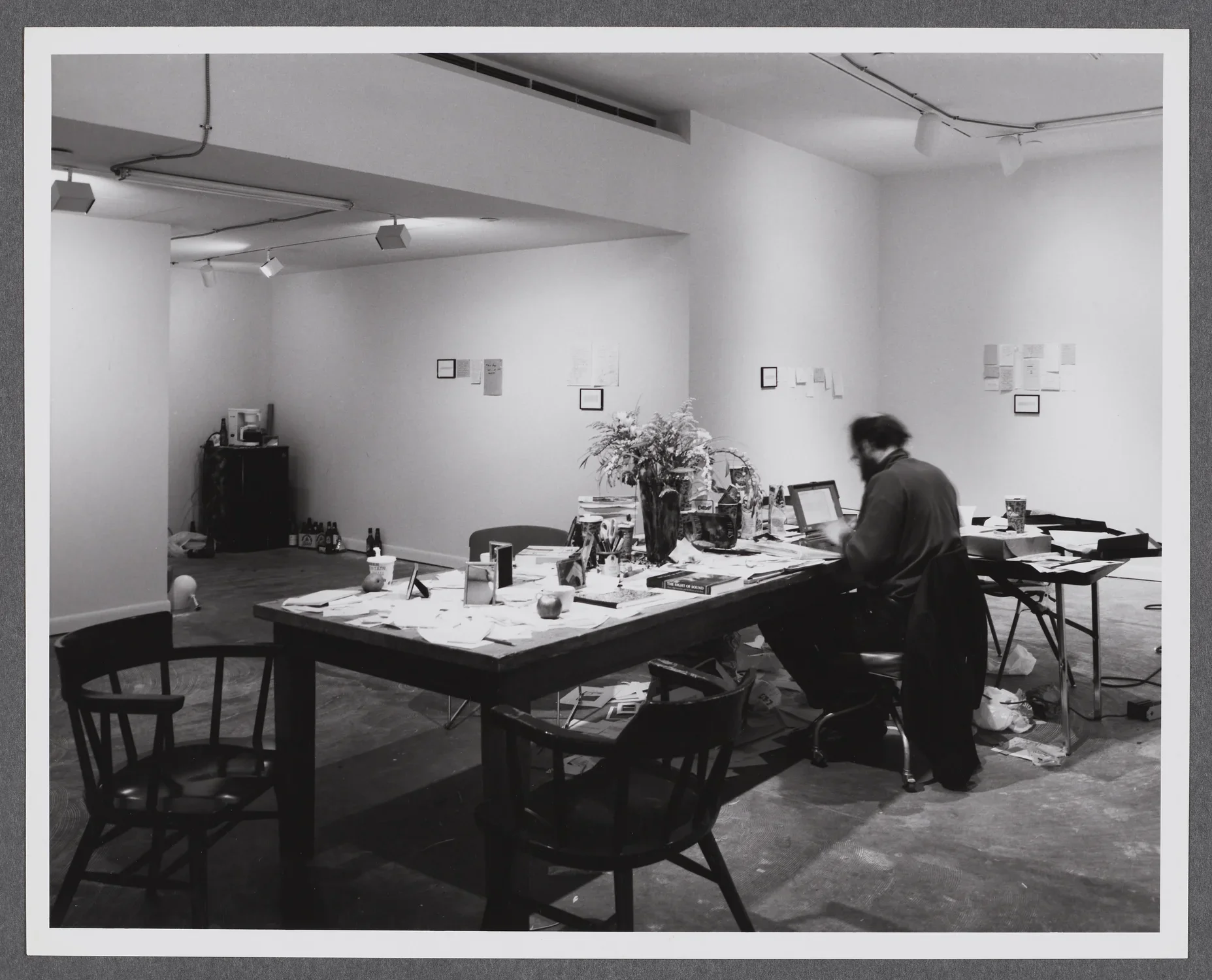 Black and white image of a man at a long table, its top strewn with paper, small miscellaneous items, and a vase of flowers.
