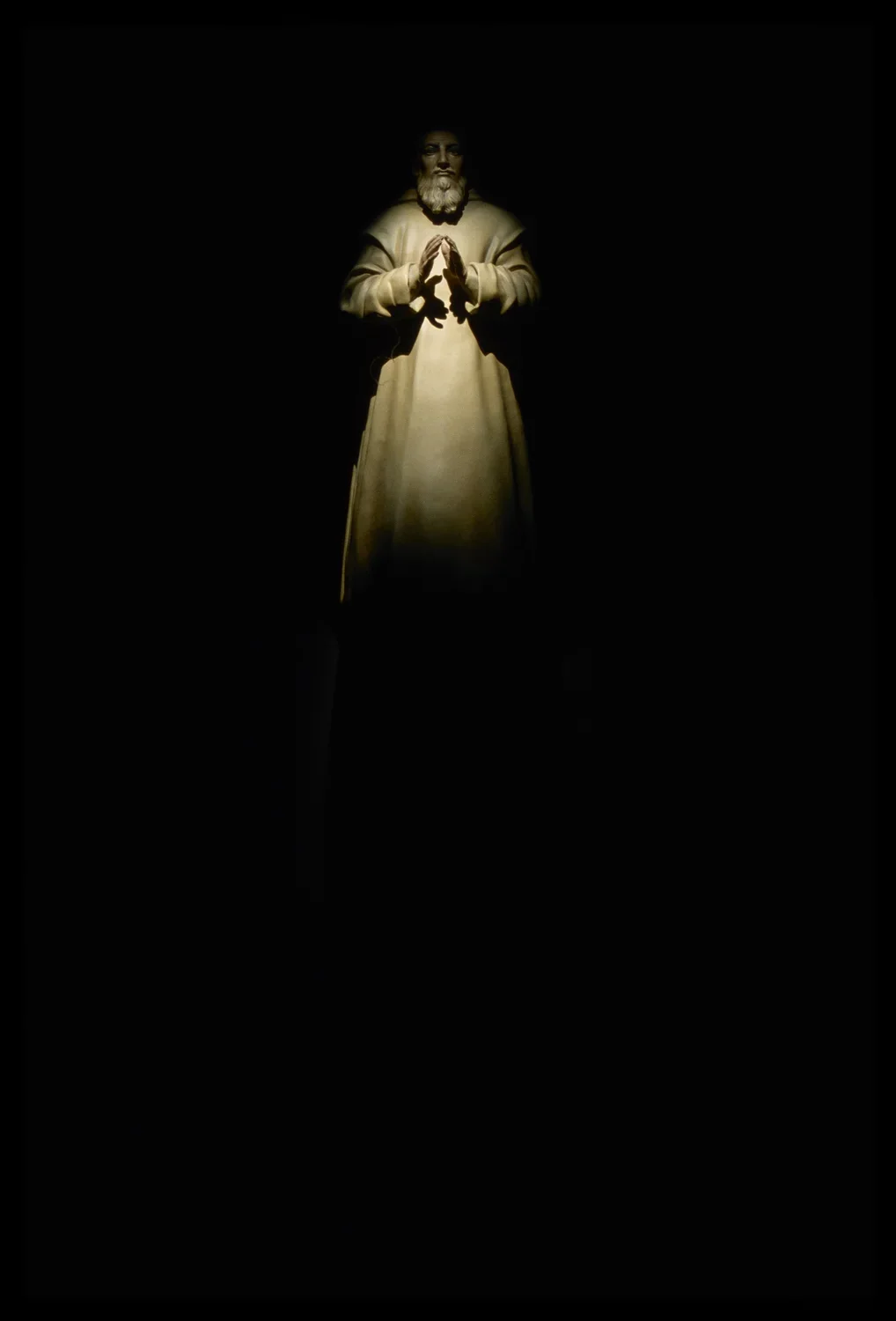 Spotlit polychromed sculpture in dark gallery room of saint in robe, with beard and hands in prayer, looking forward.