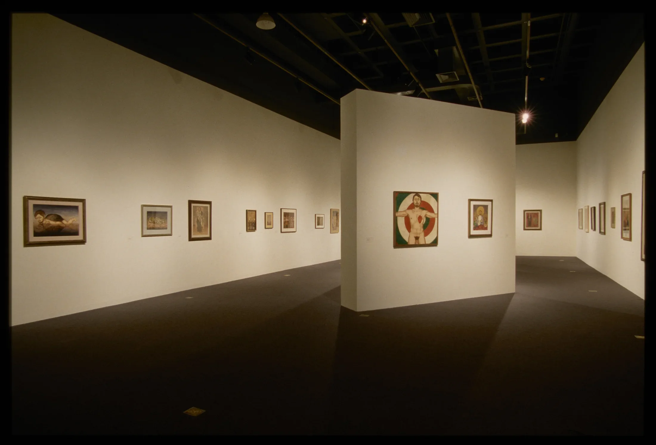 Framed paintings hang on three walls and a center partition in a gallery space.