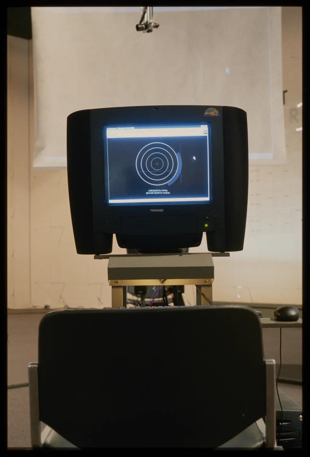 A black monitor displays concentric circles, and is mounted on a metal stand. A film screen is suspended in the background.