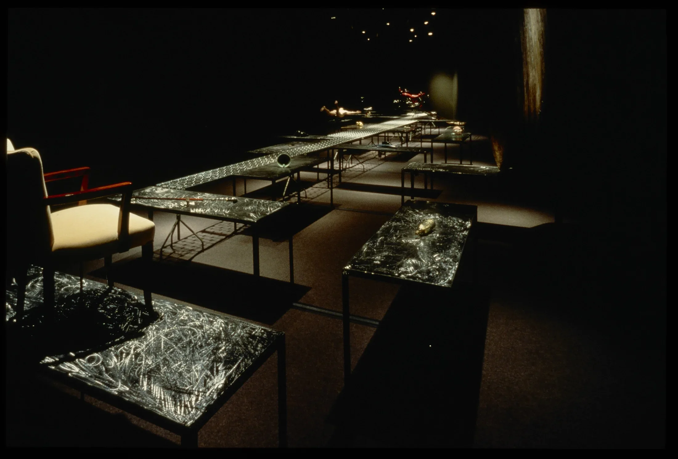 Dark room with spotlit series of black, metal, scratched tables with found items on them, with chair sitting off to the side.