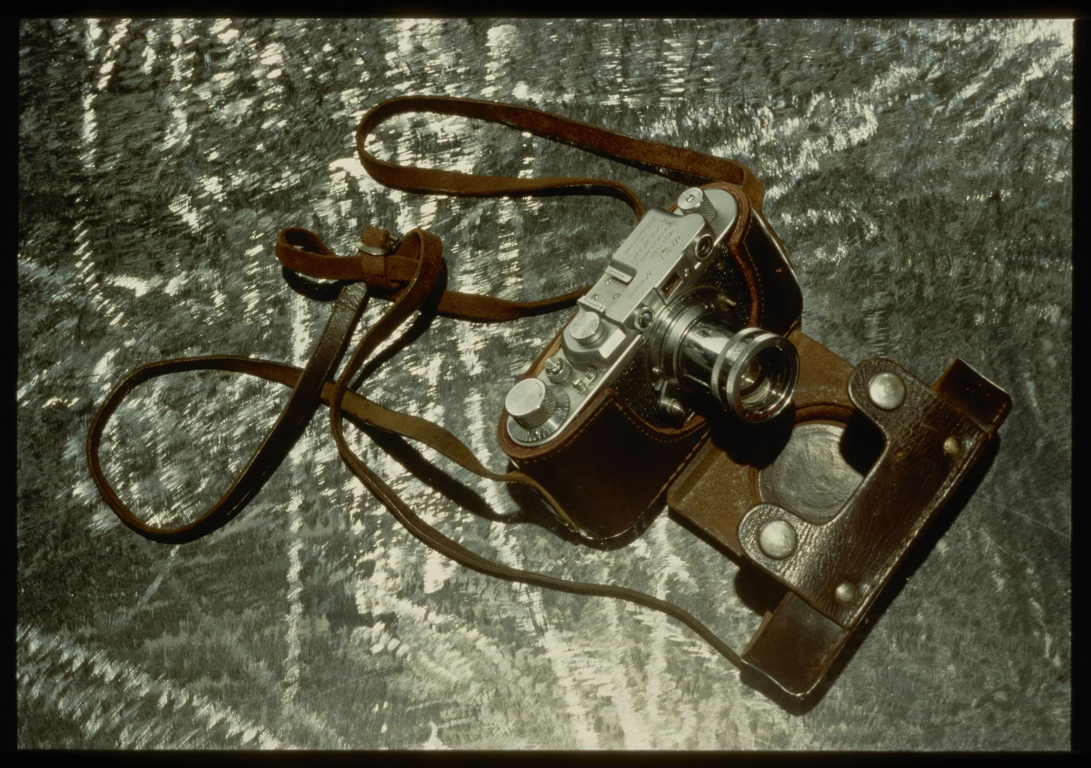 Top view of vintage camera with leather holster and strap sitting facing down right on scratched metal table.