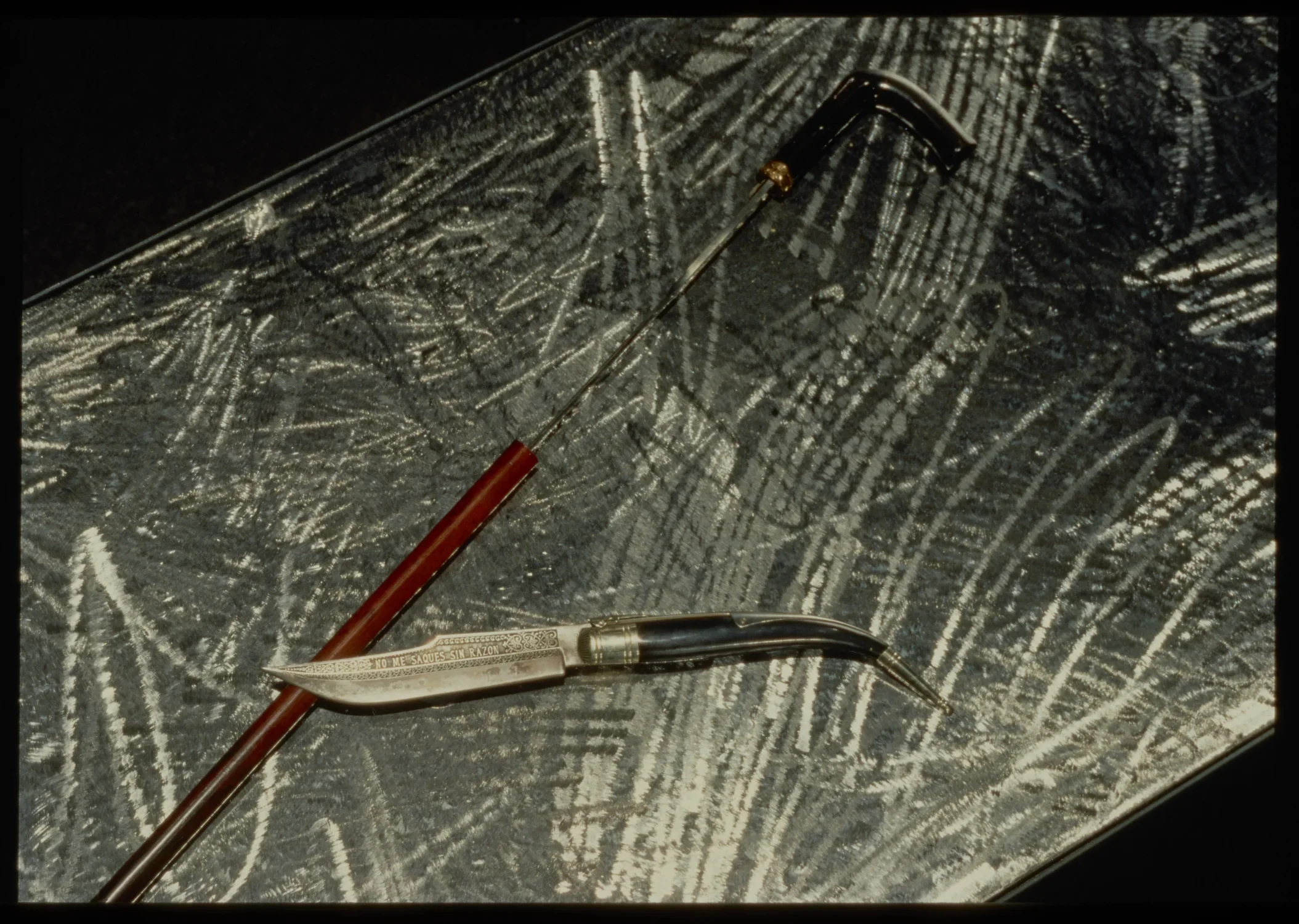 Top view of vintage knife laying on top of red wooden cane with metal handle, on top of scratched metal table.