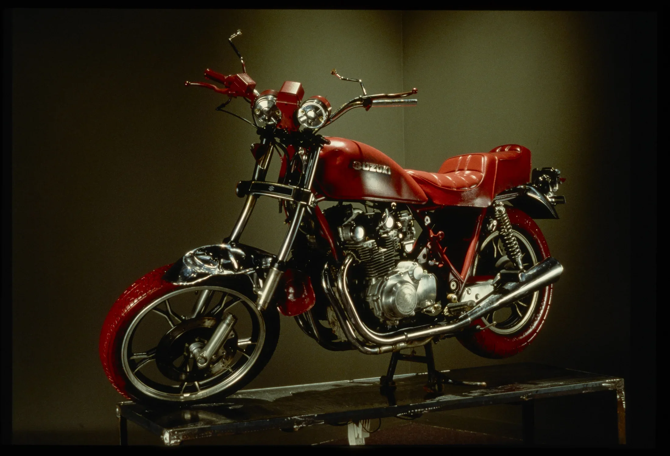 Red and chrome vintage motorcycle sitting on stand, spotlit in dark gallery.