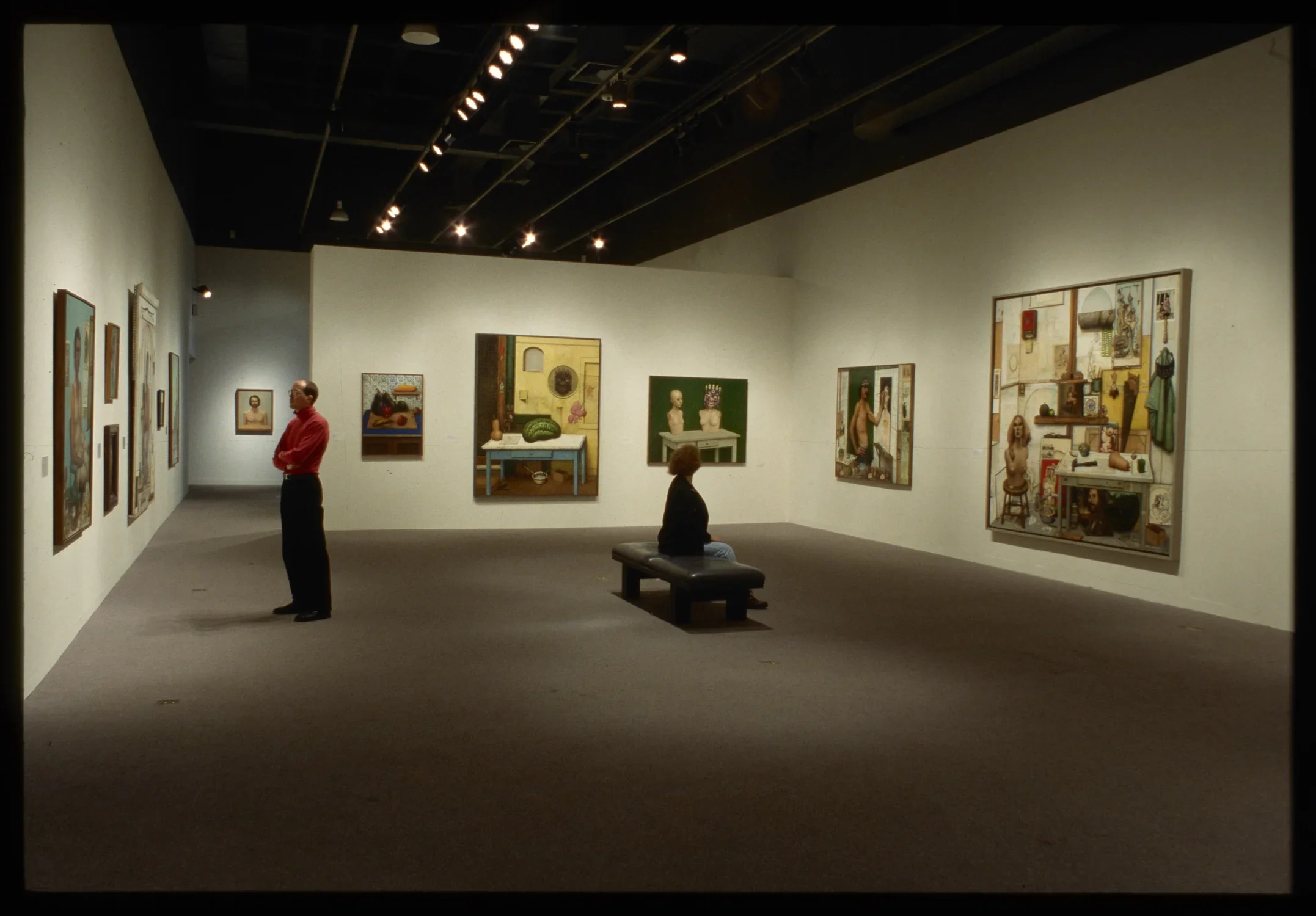 Male figure in foreground painting on left, a colorful geometric painting next to a smaller one on the back, gallery wall.