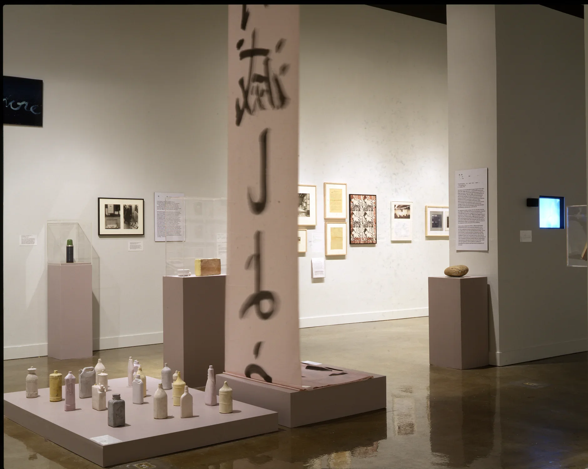 Muted colored bottles on a base, calligraphy on a large hanging scroll, art on walls and pedestals in the space beyond.
