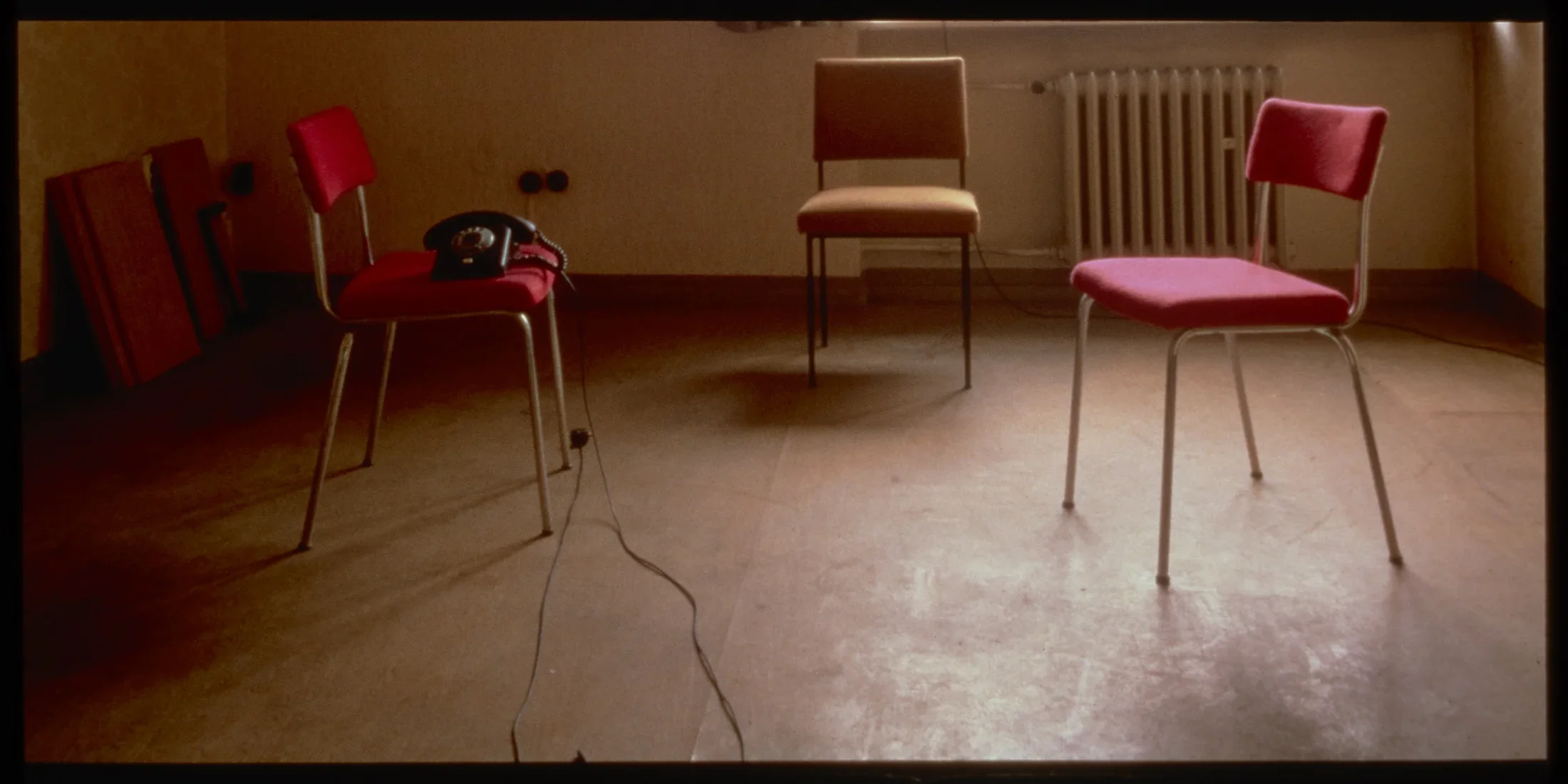 2 red chairs and 1 organe chair in an empty room with a telephone on one chair. 