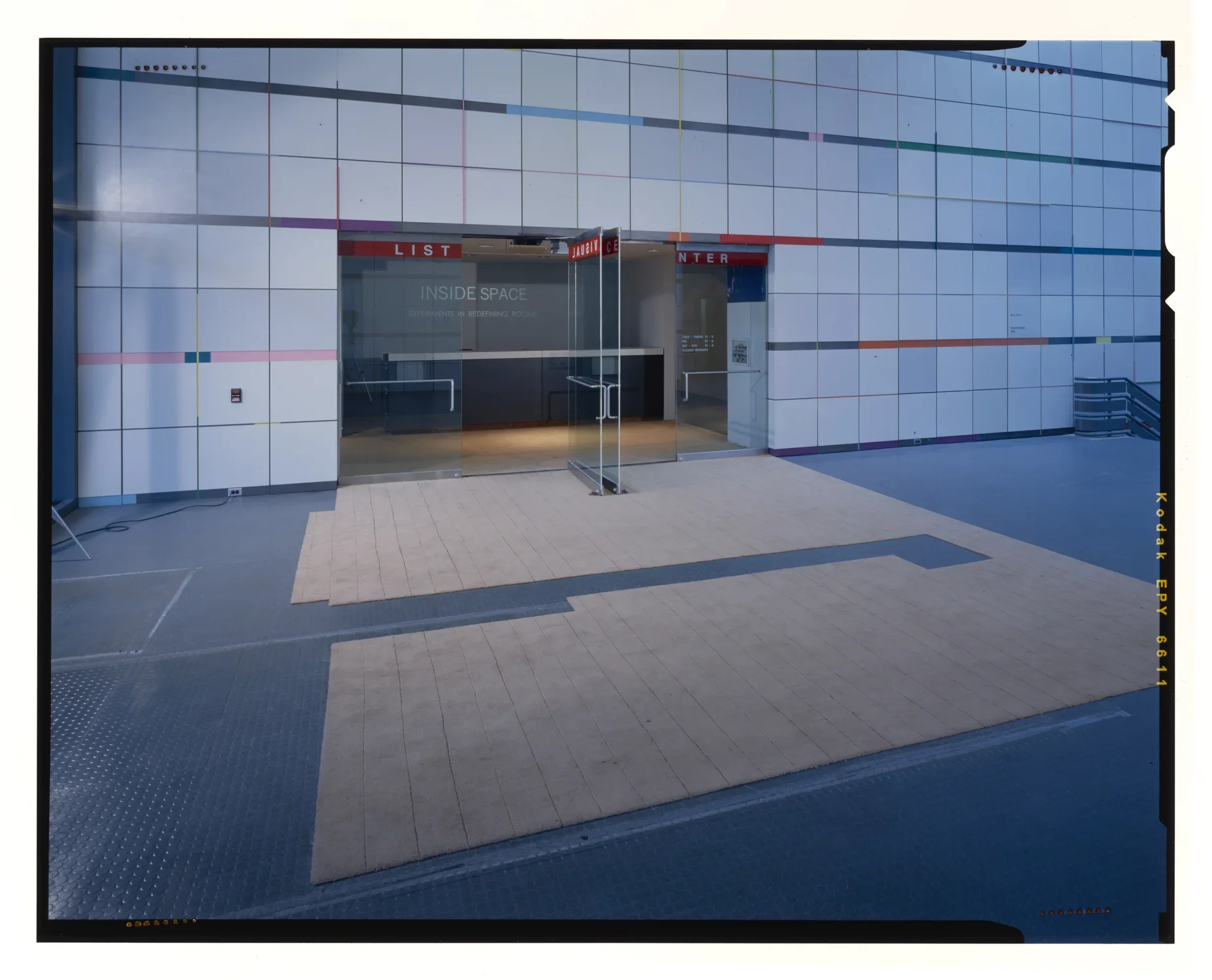 Large carpet with a long rectilinear shape cut out of the middle, lies in front of the glass door entryway of the gallery.