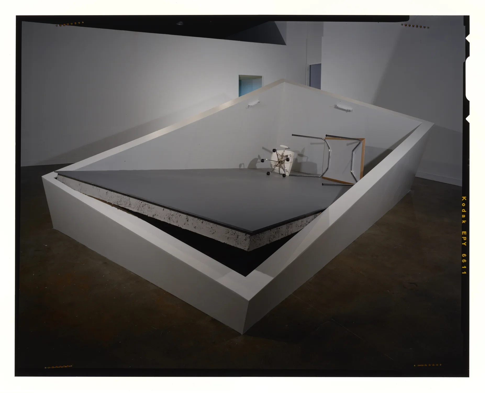 White office furniture is toppled on a grey floor that tilts up, in a distorted white room of 4 low walls that vary in height.