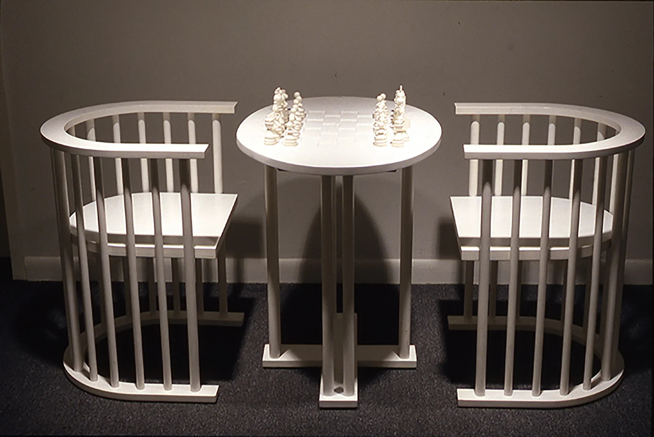 Straight on view of a wooden chess table with pieces set up and 2 wooden chairs, all painted white