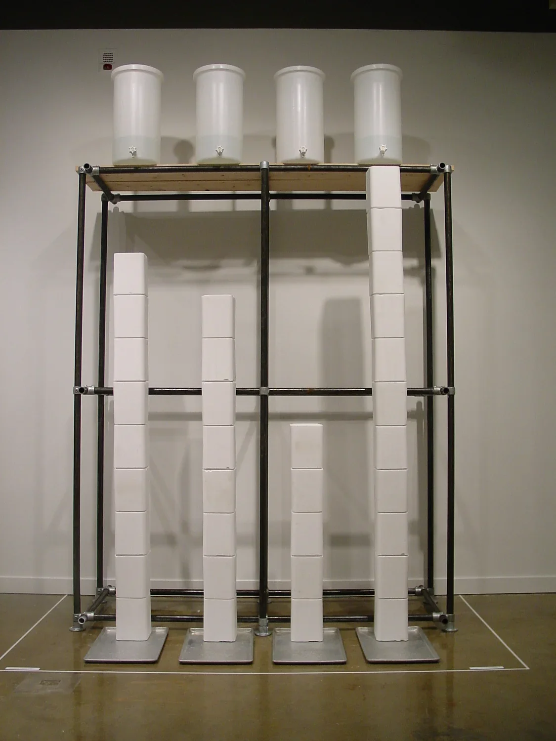 White compressed salt blocks stack top 4 aluminum trays. Behind, a black framed structure is against the wall