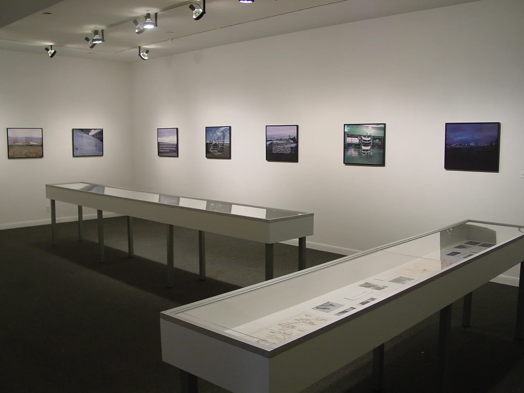 Small square artworks are spaced evenly along two walls with 2 lengthy vitrines, perpendicular to each other
