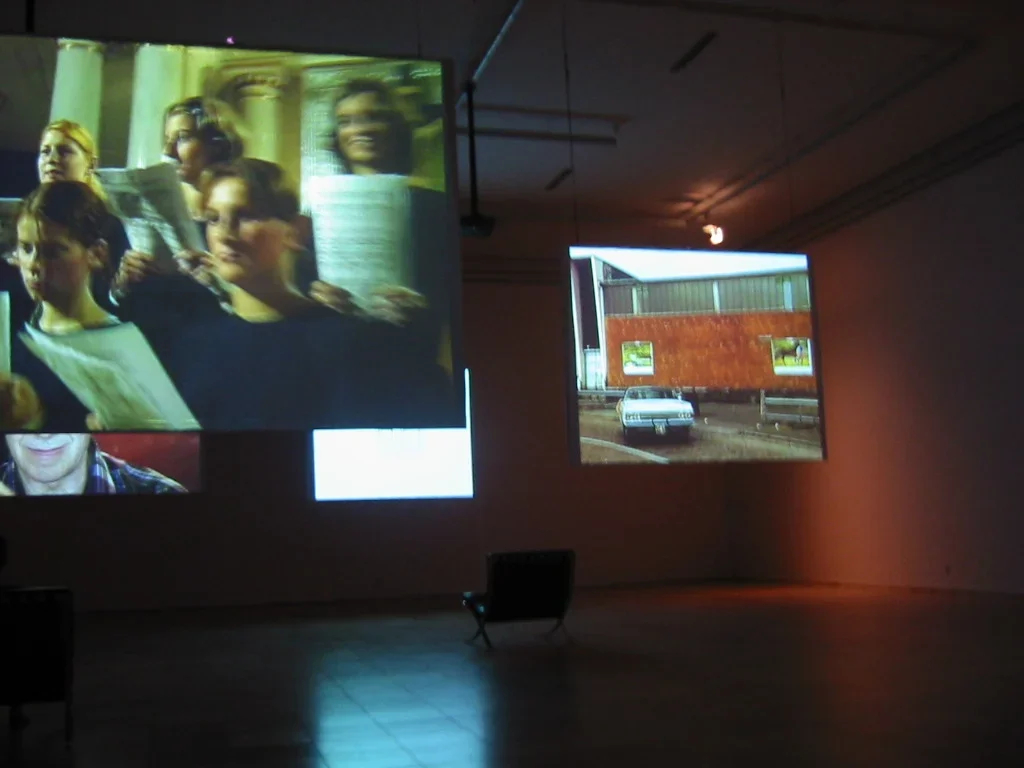 4 projection screens hang down from the ceiling in a dark room. One single chair sits empty in the background