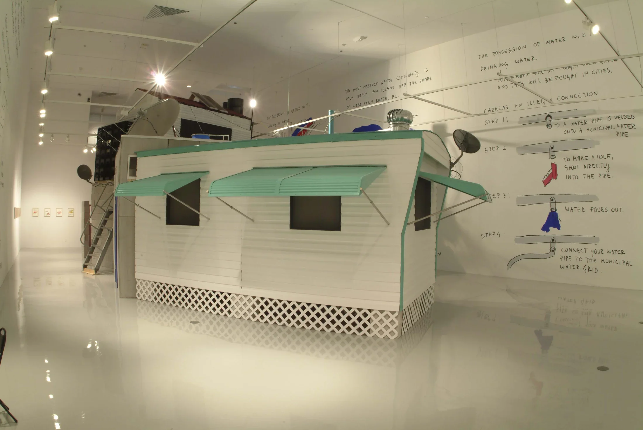 A small white house with turquoise awnings sits in gallery space with writings on the wall behind