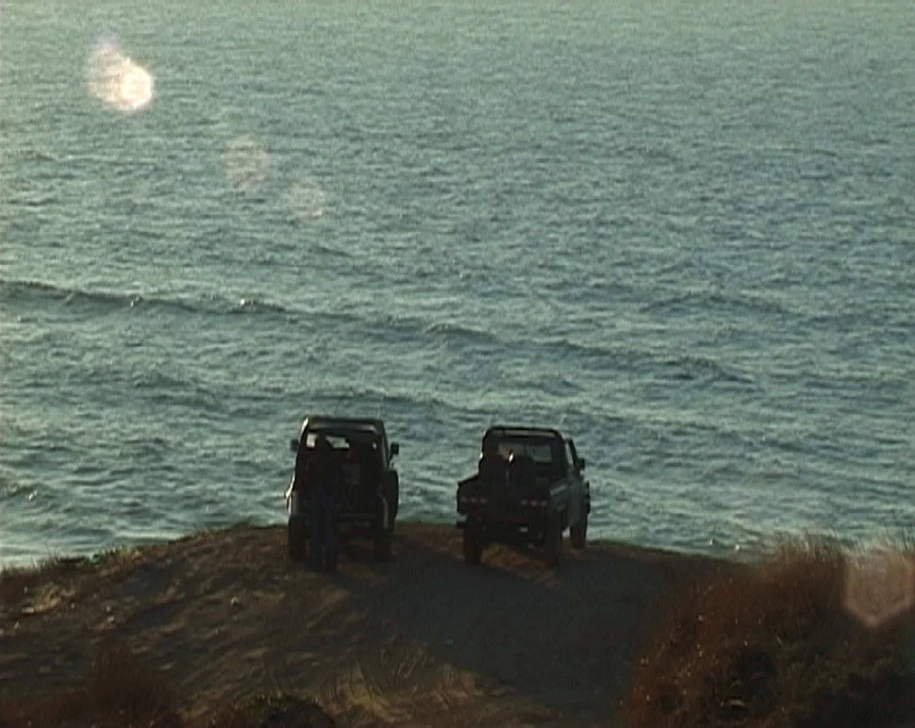 Two off road vehicles are parked at the edge of a cliff overlooking an ocean, a person stands at the rear of one of them.