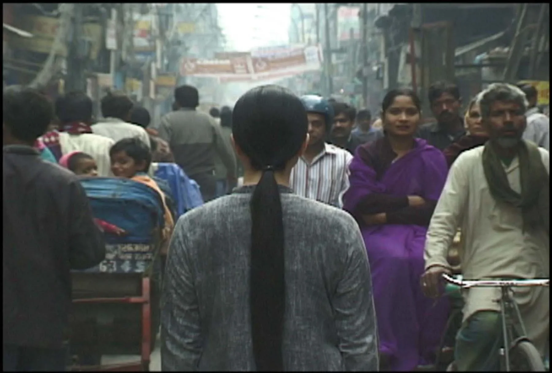 A woman with long black hair dressed in gray clothing stands with her back to the camera on a crowded street in Delhi.