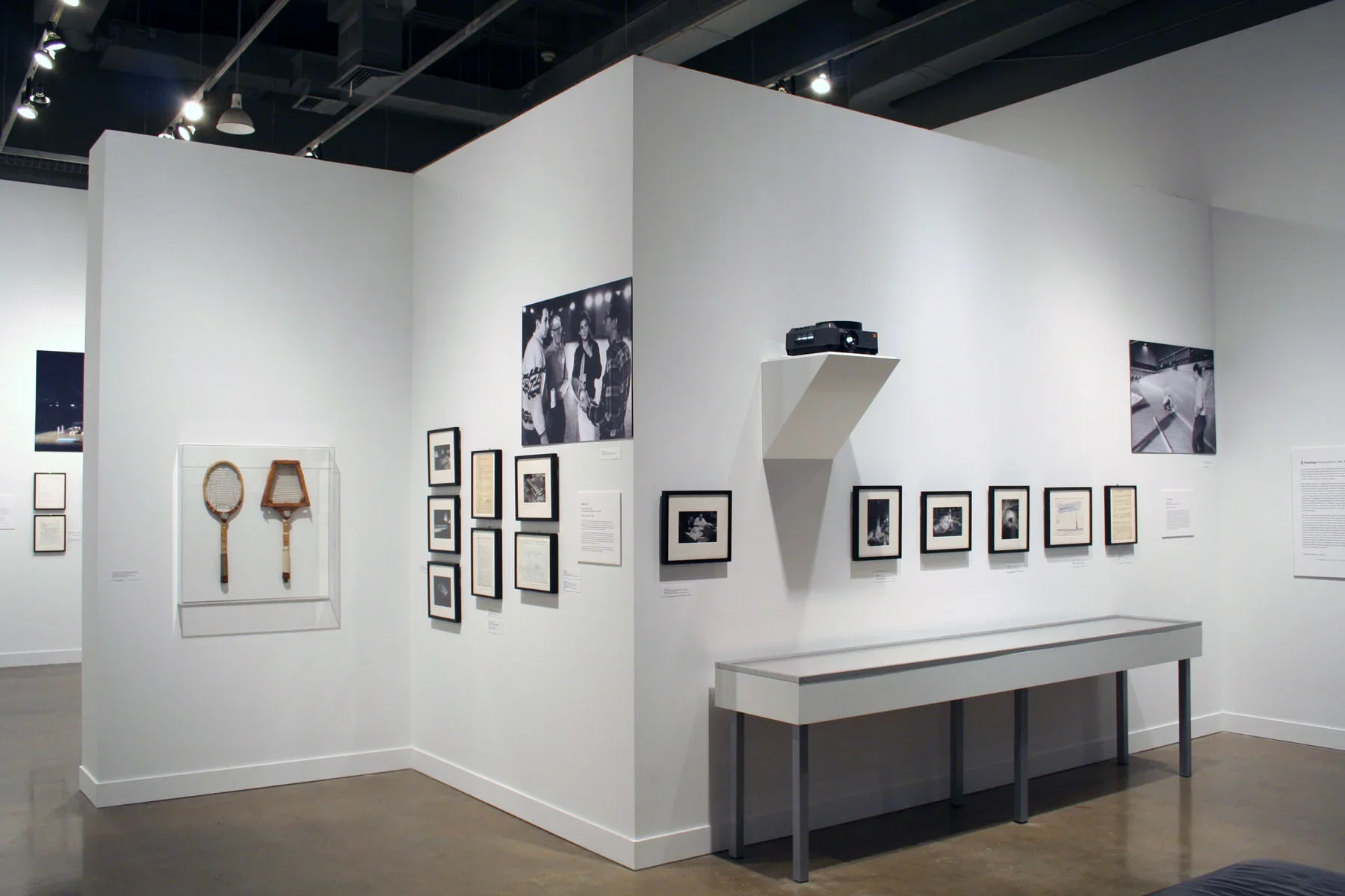 View of gallery shows 2 tennis rackets in a case, framed works and large photos mounted on walls and a glass top display case.