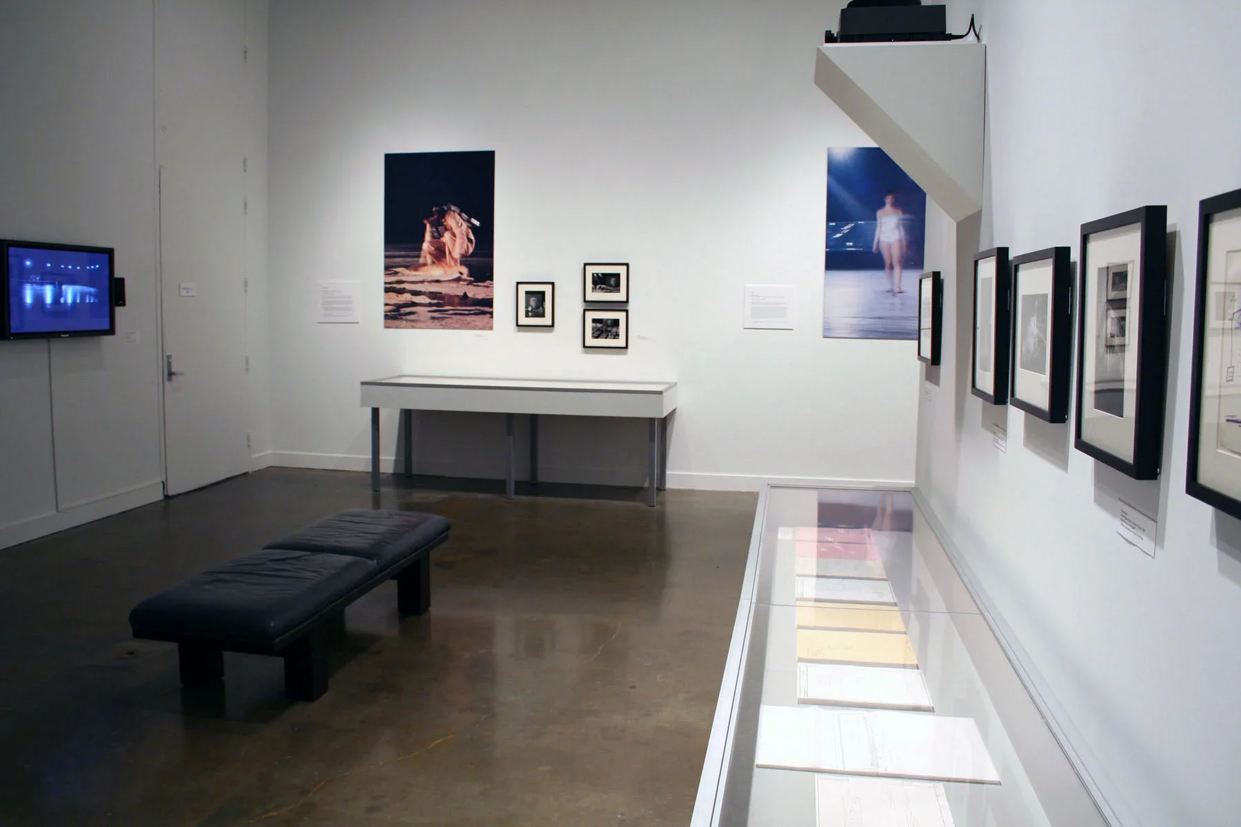 Miscellaneous framed photographs hang against a white gallery wall while paper artifacts sit in a glass vitrine.