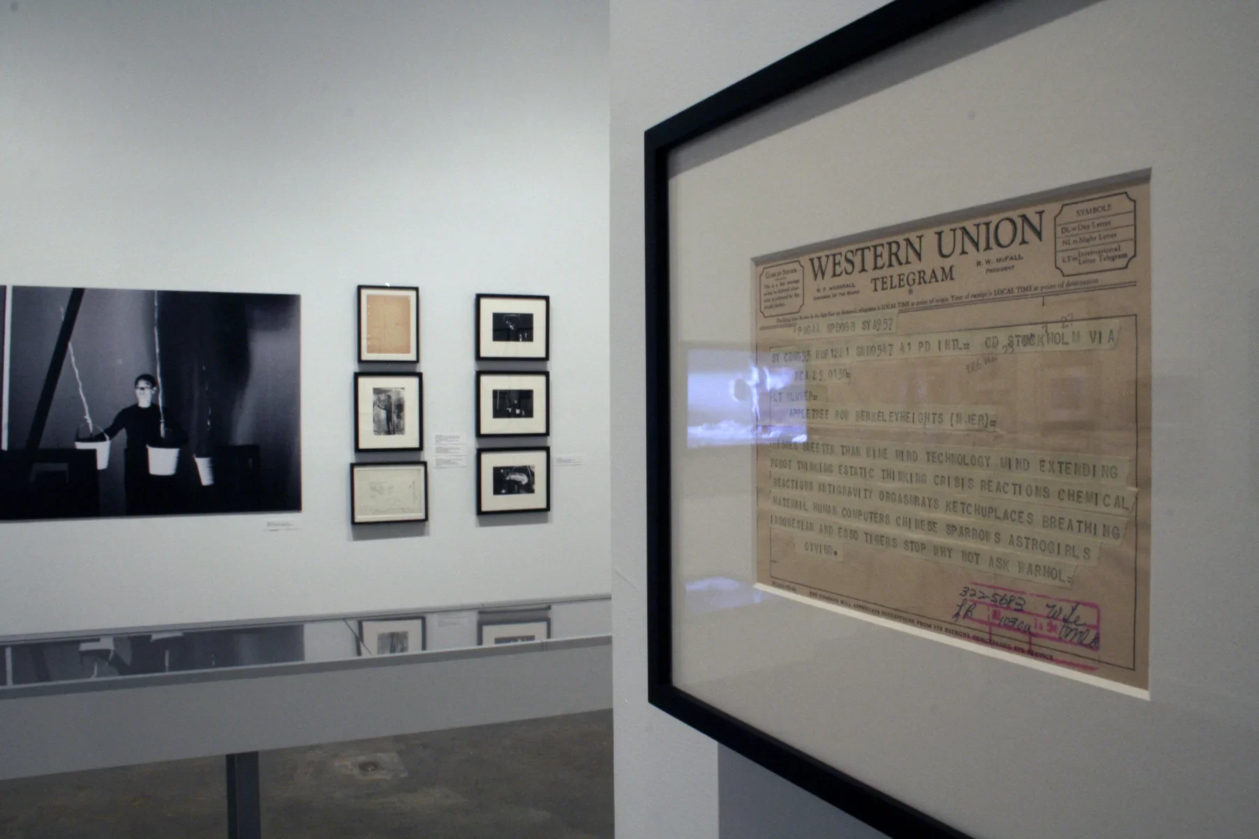 View of gallery with framed artworks and photos hung on walls, a long display case, and a video monitor with a bench in front.