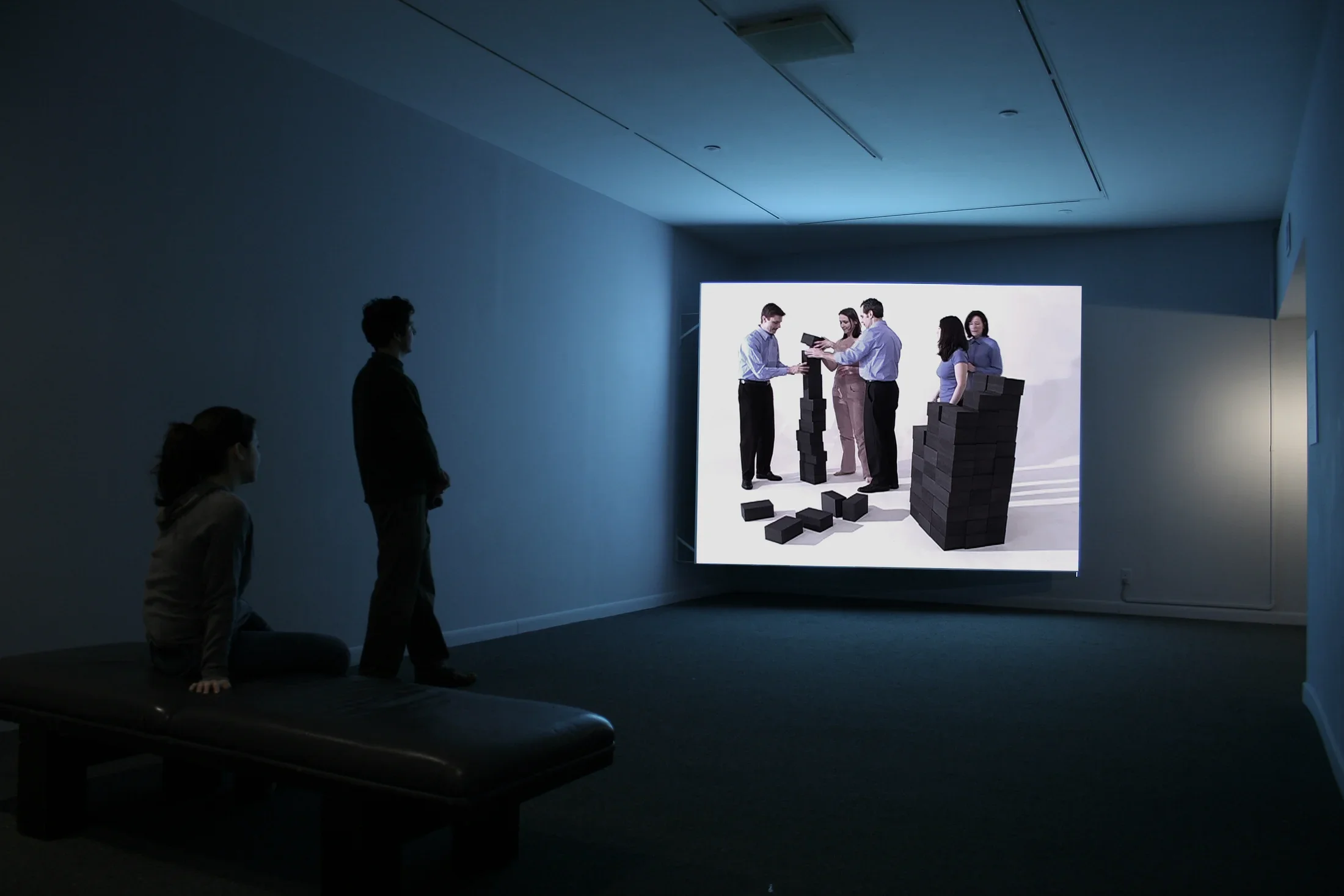 2 viewers looking at a large screen with 5 people working with blocks.