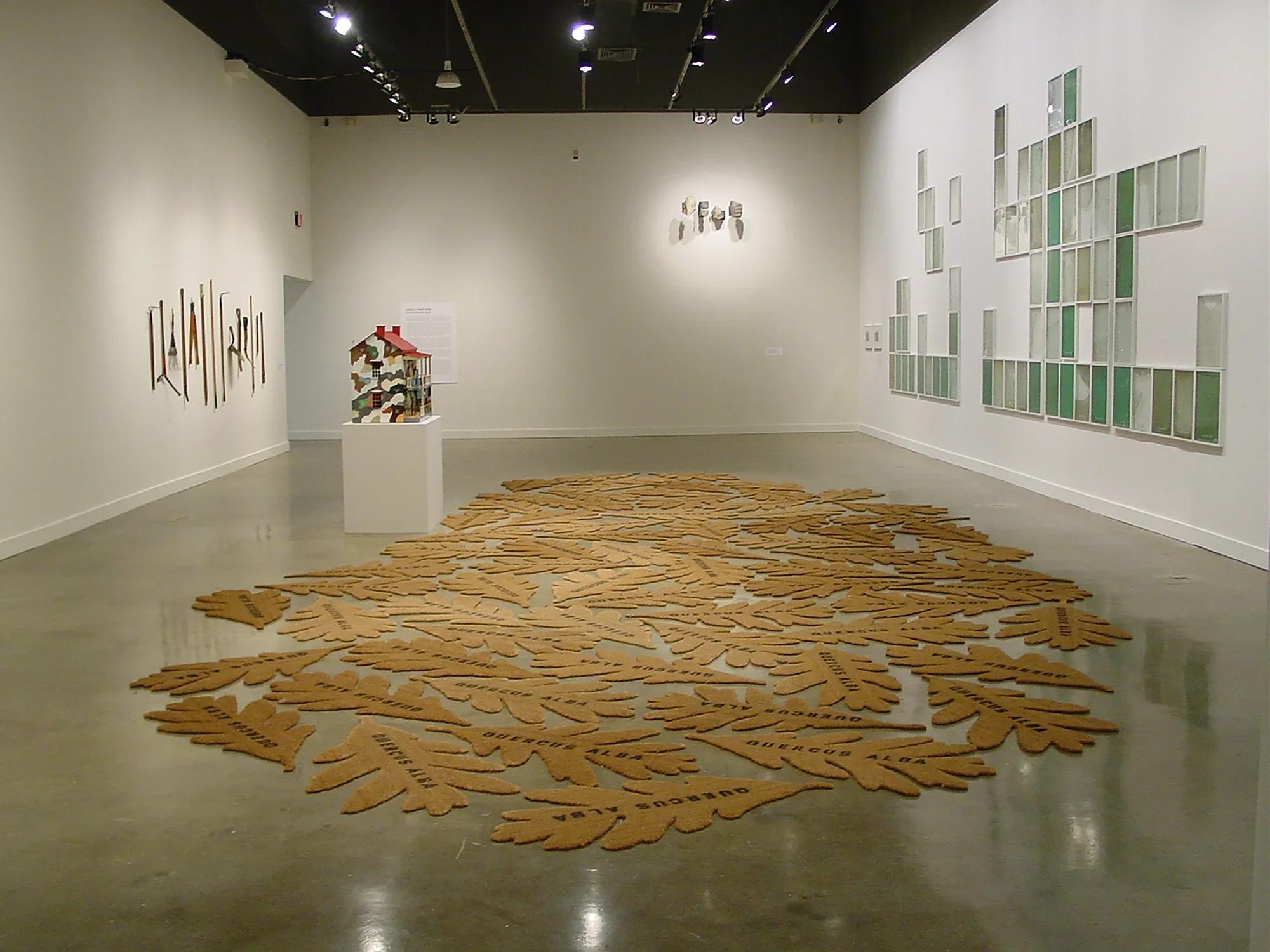 Multitude of oak leaf shaped mats beside a colorful model house sculpture on pedestal and 3 artworks on surrounding walls.