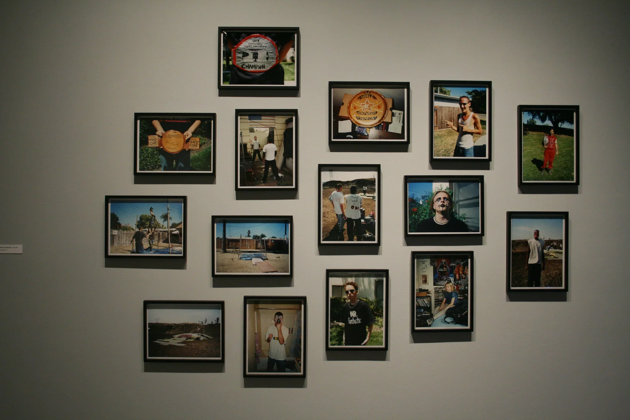 15 color photographs of young men, objects and landscapes in black frames hang on a wall in an abstract grouping.