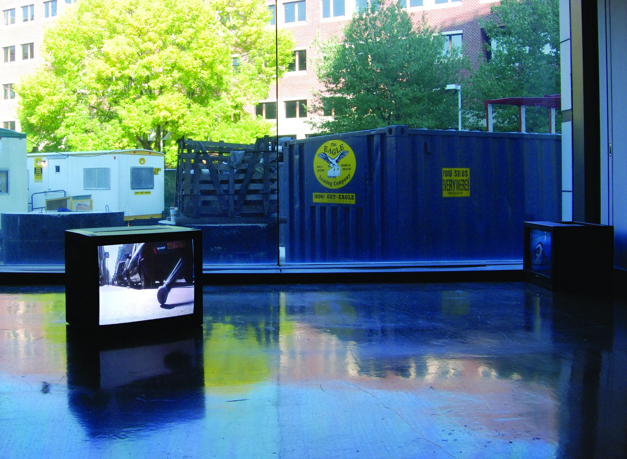 In front of a picture window looking out on construction, two monitors sit on the floor, one with a view of cars on a street
