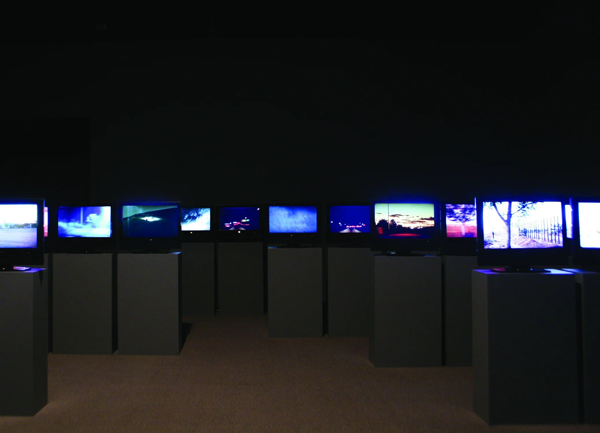 Many screens installed on podiums in a darkened gallery. 